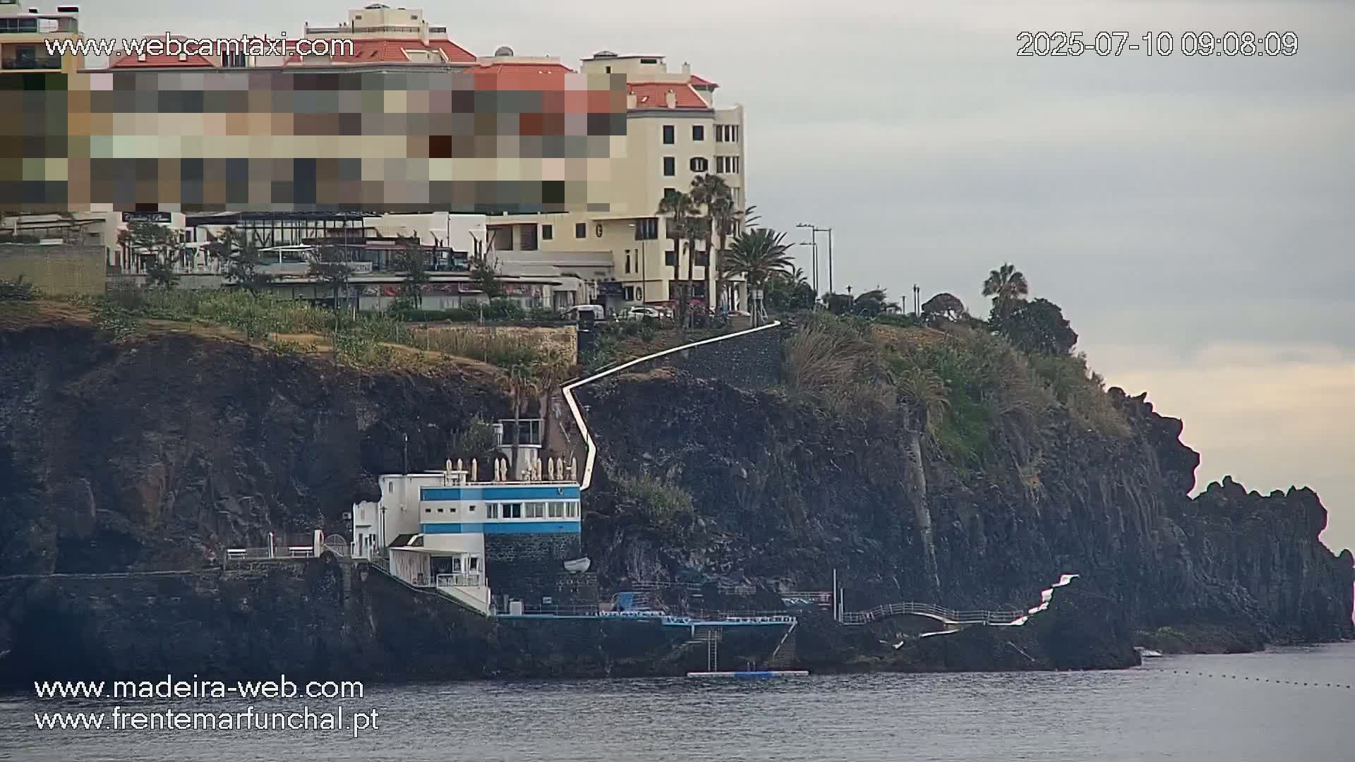 A cliffside building with a small structure at its base overlooks calm ocean water under an overcast sky.