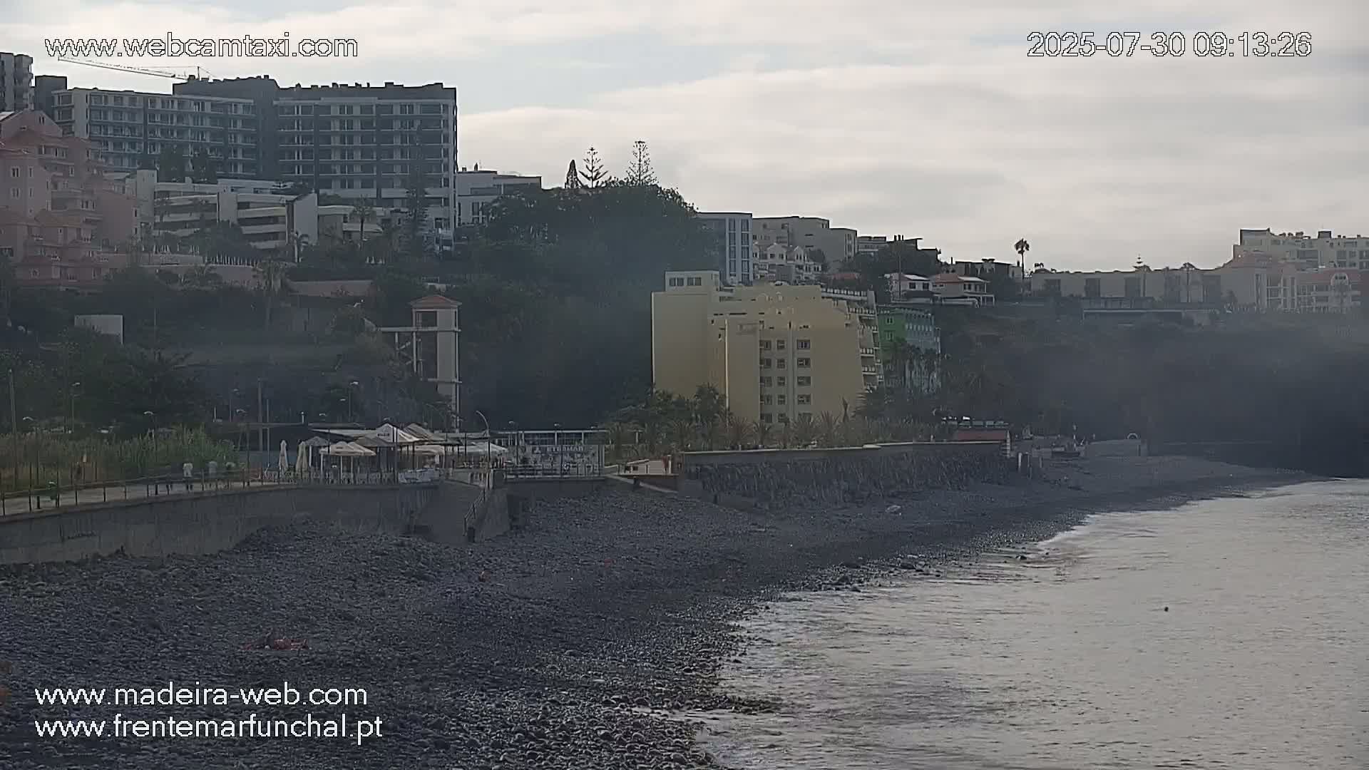 A dark-colored, pebble beach meets a calm ocean under an overcast sky, with multi-story buildings and lush vegetation situated on a cliff overlooking the shore.