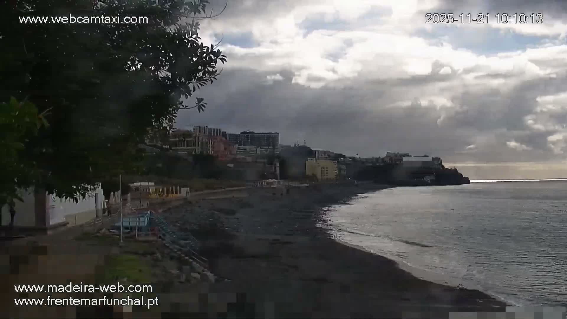 A coastal view on a day with heavy, broken clouds and bright patches features a dark pebble beach with gentle waves, a city built on hills in the background, and a headland extending into the calm sea.