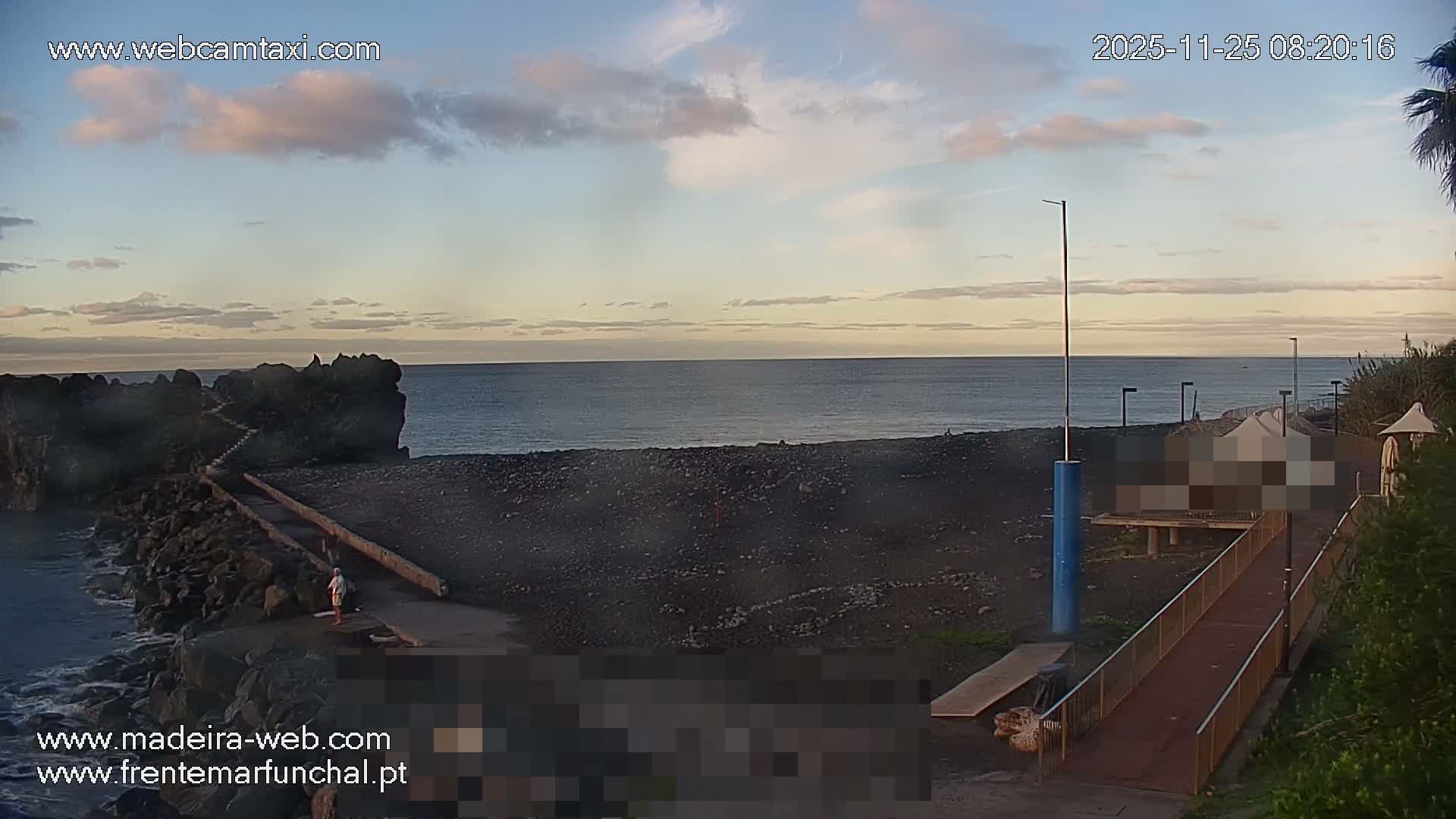 A tranquil coastal scene unfolds under a partially cloudy sky with calm waters, showing a dark pebble beach, a rocky breakwater with a lone figure, and a paved seaside path with railings and greenery.