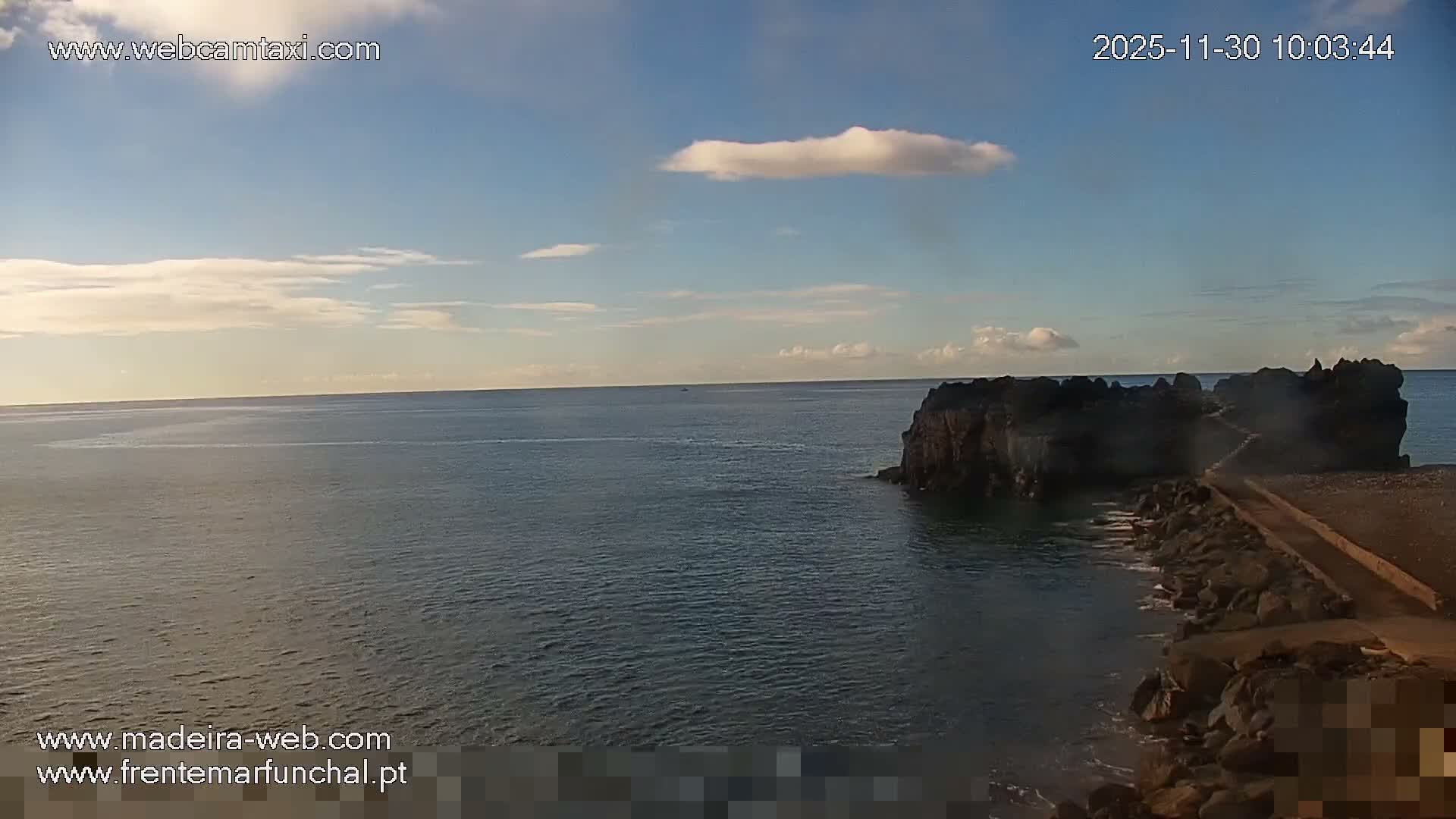 A tranquil coastal scene features a calm blue sea under a partly cloudy sky, with a dark, rugged rocky formation on the right connected to the shore by a pathway.
