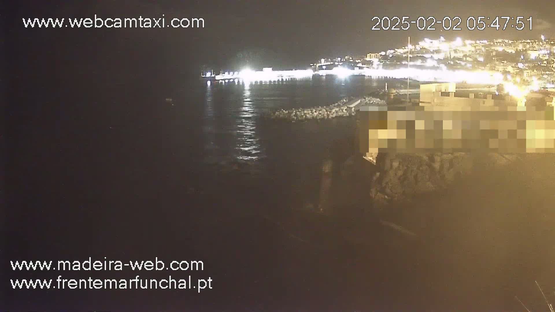 A nighttime, seaside view shows a calm body of water lapping against a rocky shore, with a brightly lit town visible in the distance.