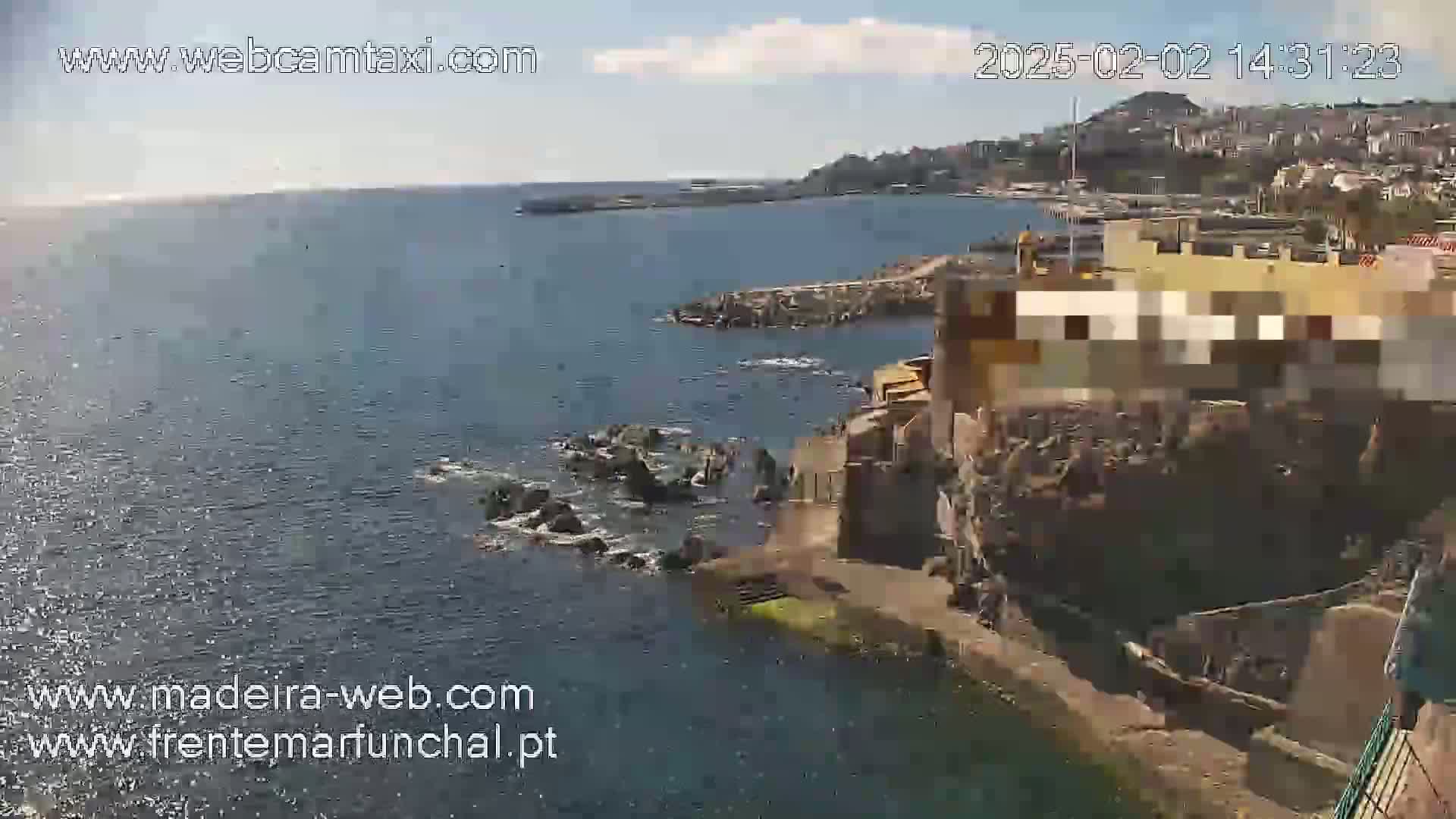 An aerial view shows a rocky coastline with a partially obscured building, a calm ocean, and a town visible in the distance under a sunny sky.
