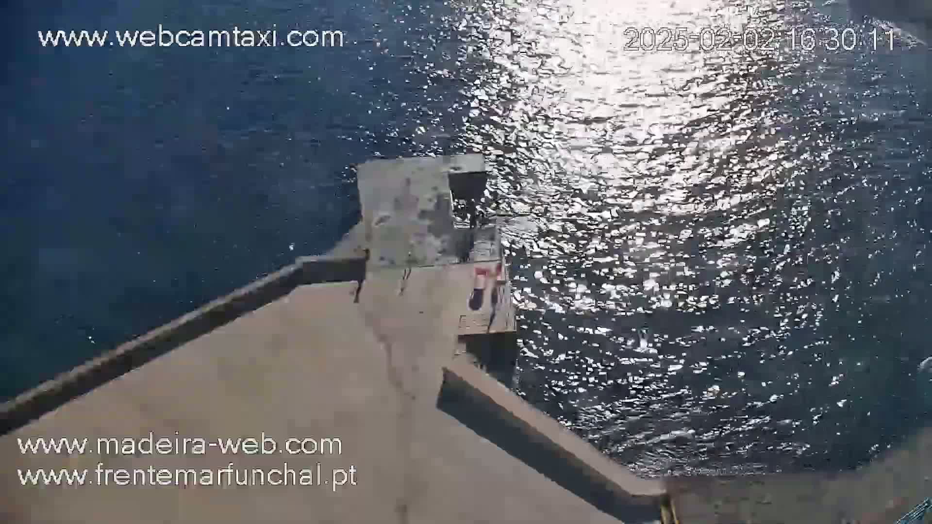 An aerial view shows a concrete platform extending into a body of dark-blue water sparkling in the sunlight.