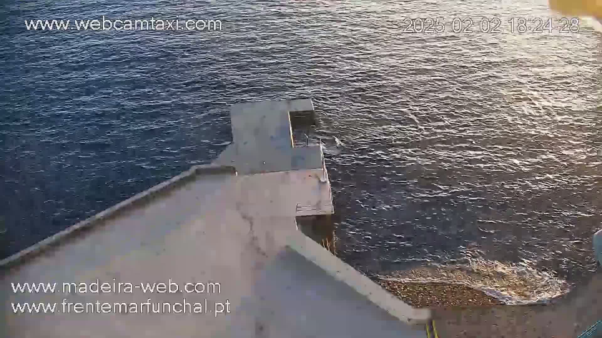 A high-angle view shows a concrete platform extending over a dark-blue body of water with small waves under a seemingly calm sky.