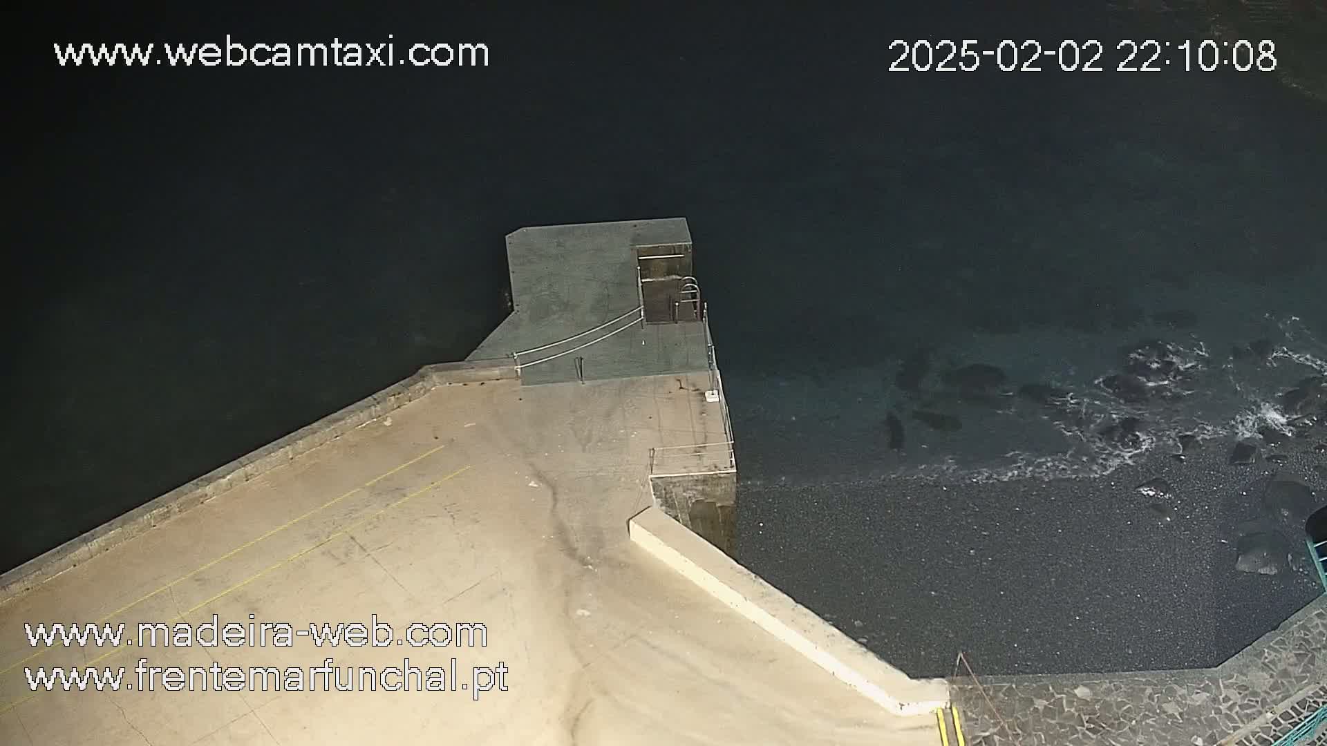 An overhead nighttime view shows a dark-colored body of water lapping against a concrete structure and a dark pebble beach.