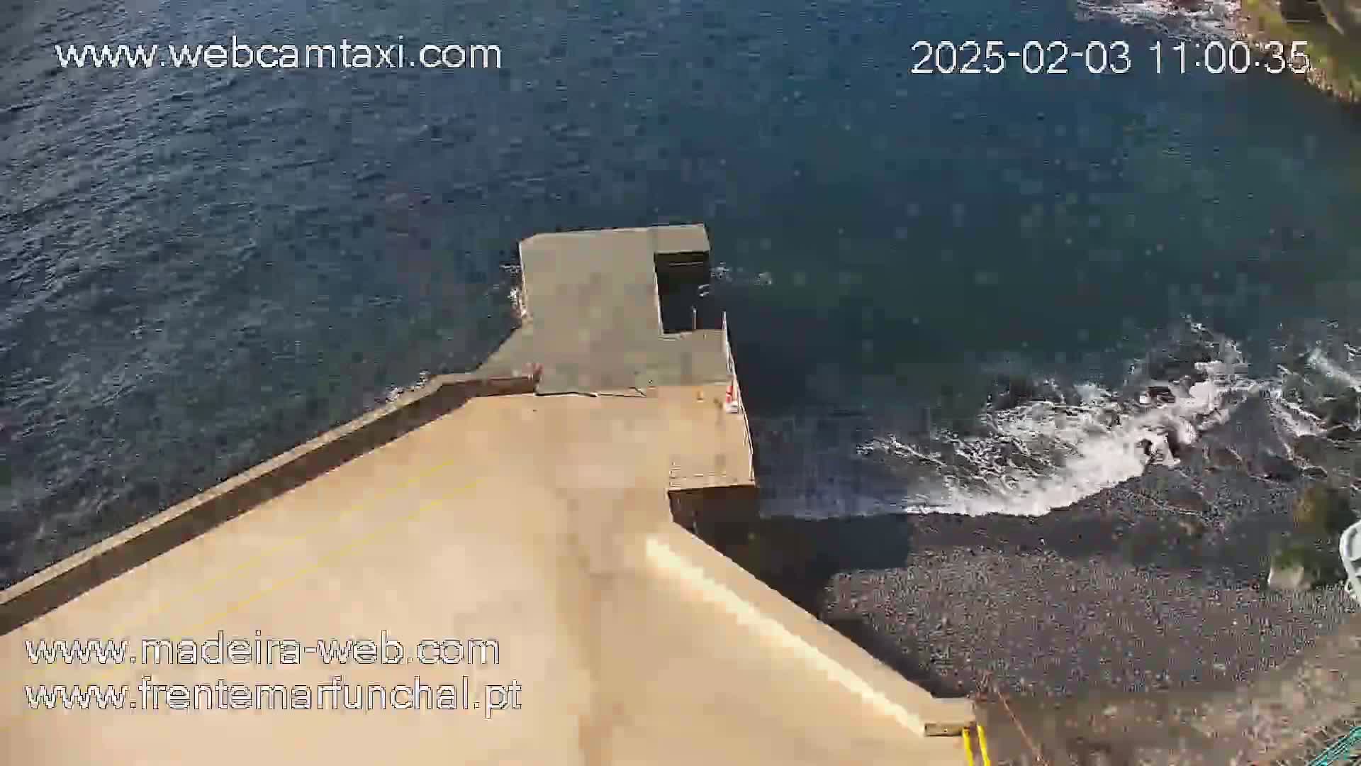 A high-angle view shows a concrete structure extending from a rocky shoreline into dark-blue water with small waves.