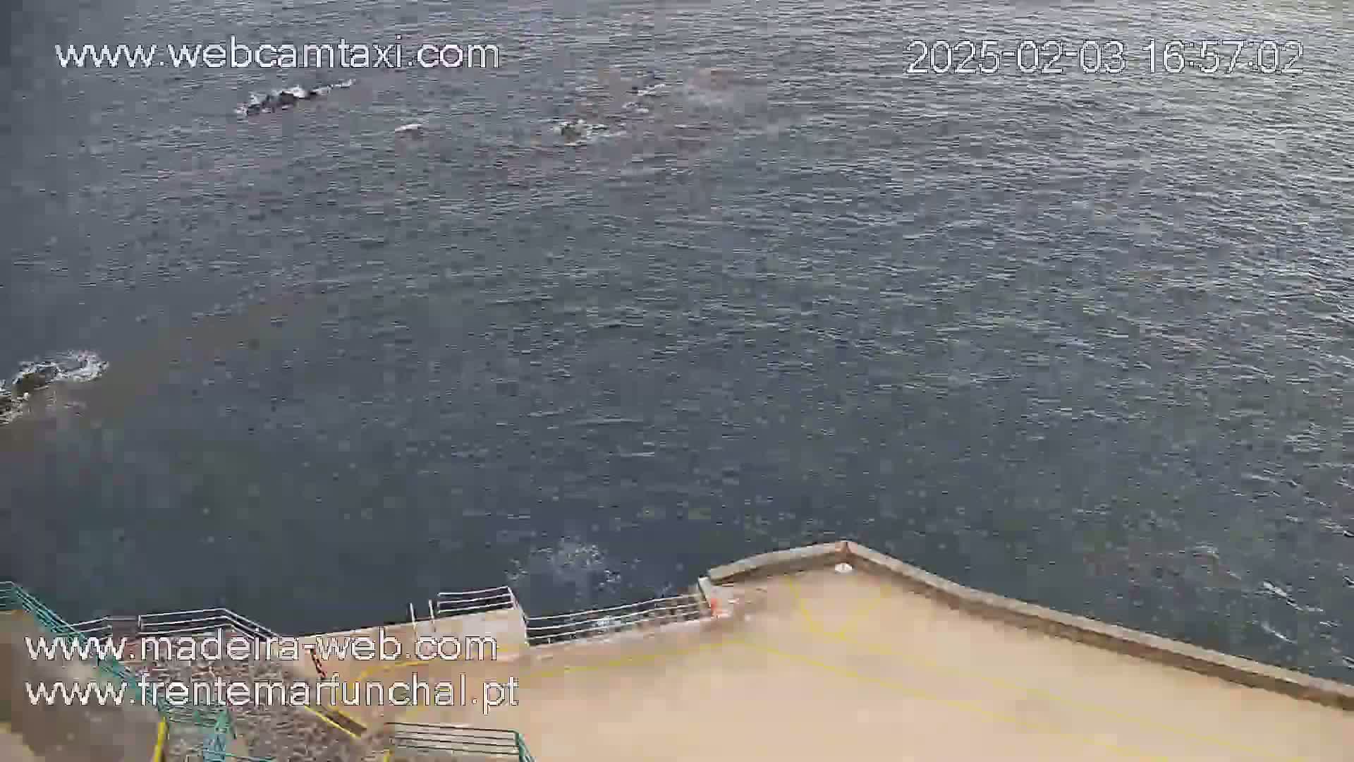 A body of dark, somewhat choppy water is seen from an elevated vantage point, next to a tan-colored paved area with metal railings.