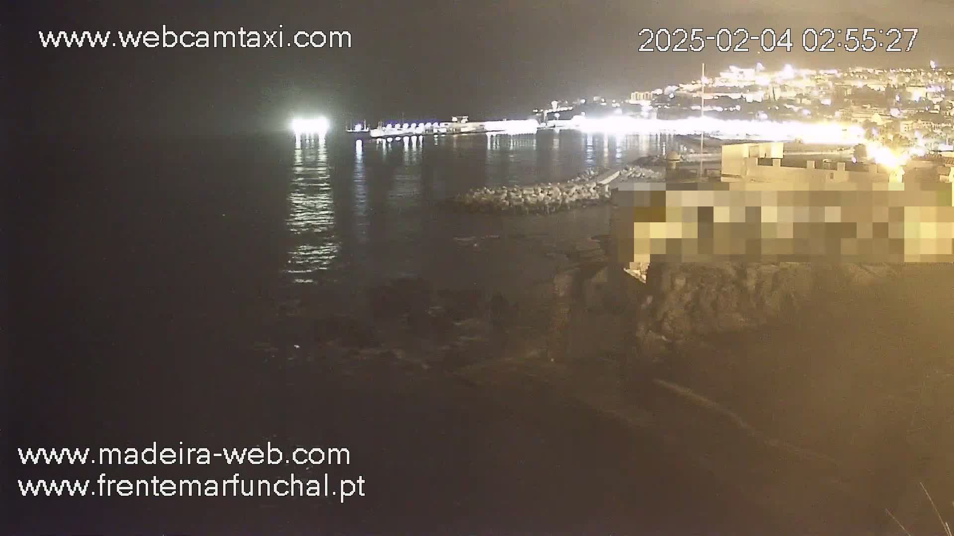 A nighttime view of a calm body of water reflecting city lights, with a shoreline featuring dark rocks and a building.
