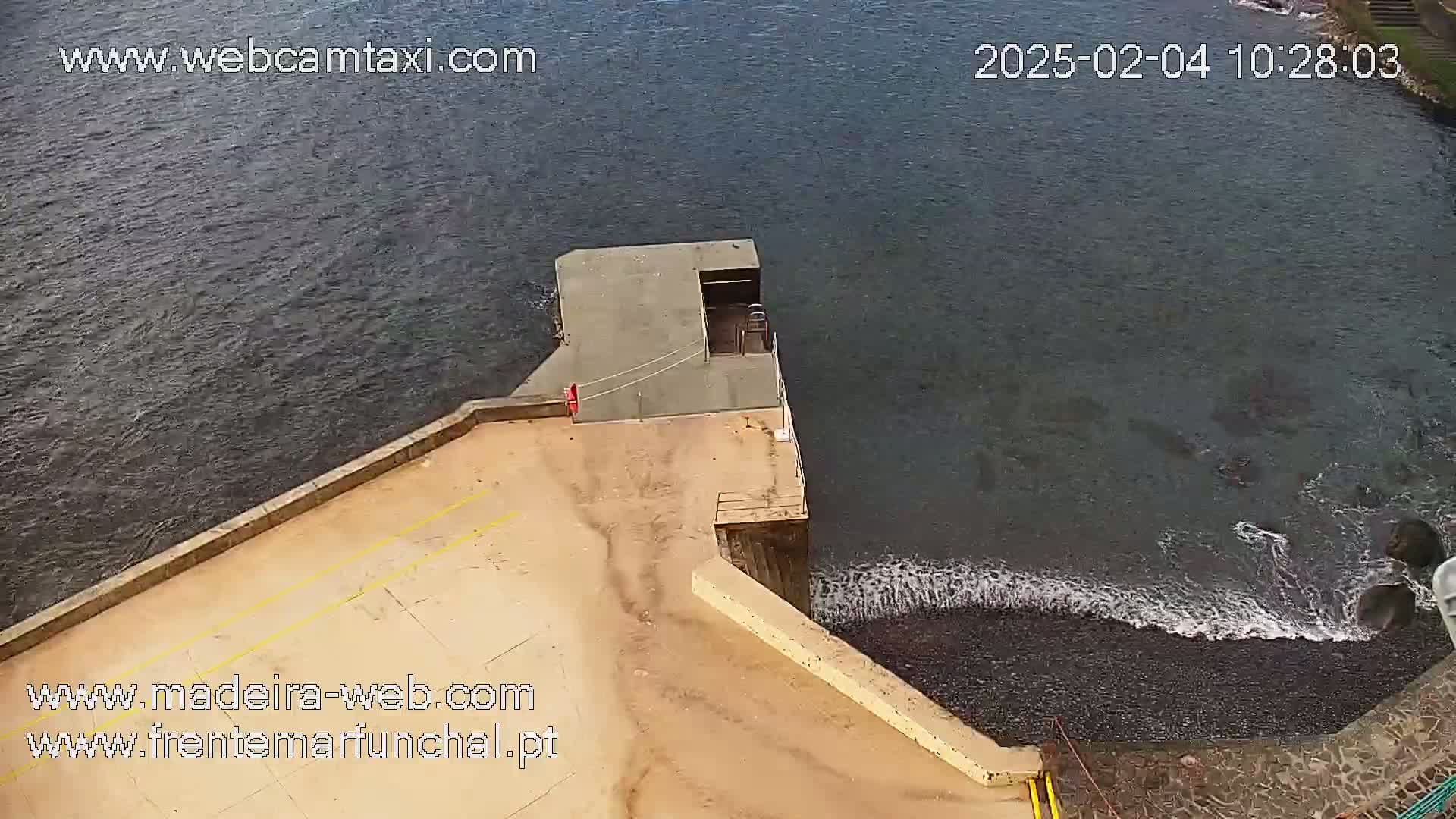 An aerial view shows a concrete platform extending into dark water with small waves lapping against a dark-colored shore.