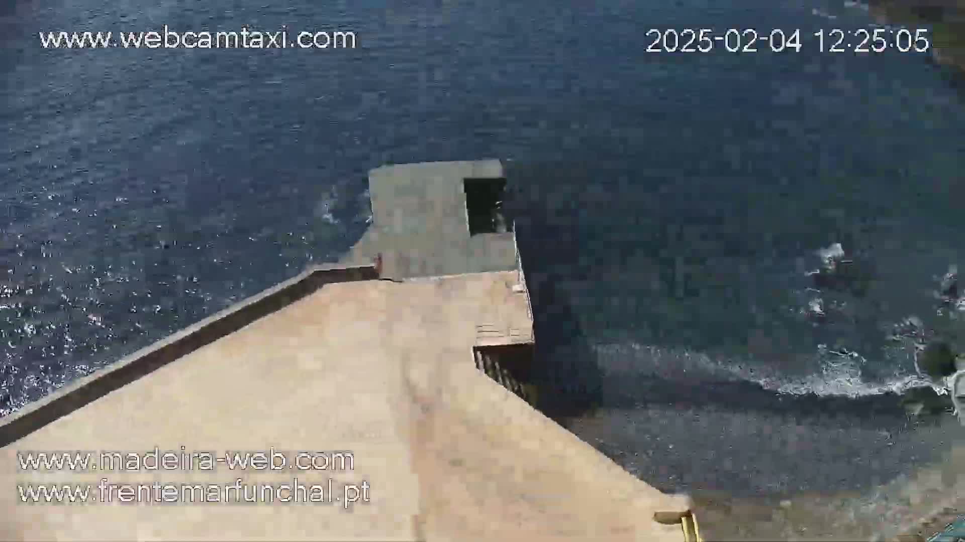 An aerial view shows a light tan concrete structure extending into dark-blue, slightly wavy water.