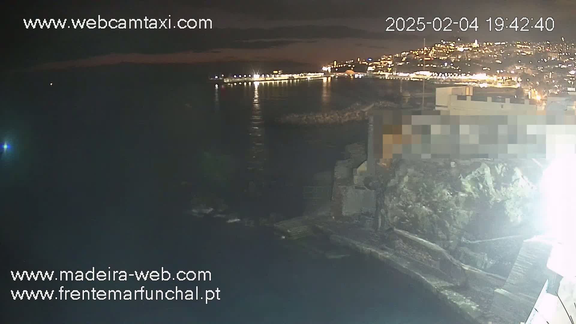 A nighttime view of a city's illuminated waterfront from a rocky vantage point overlooking dark water.