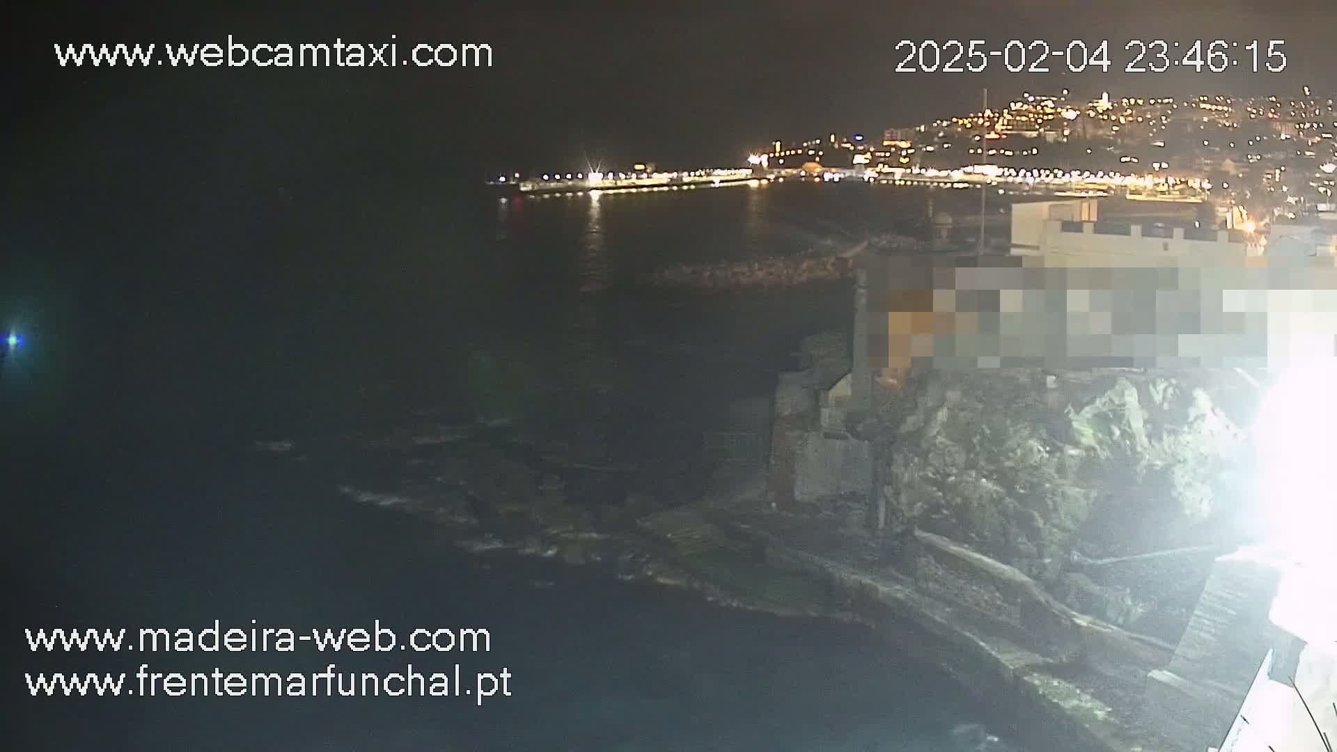 A nighttime view of a rocky coastline with a city's lights illuminating the water and a building partially obscured on the rocks.