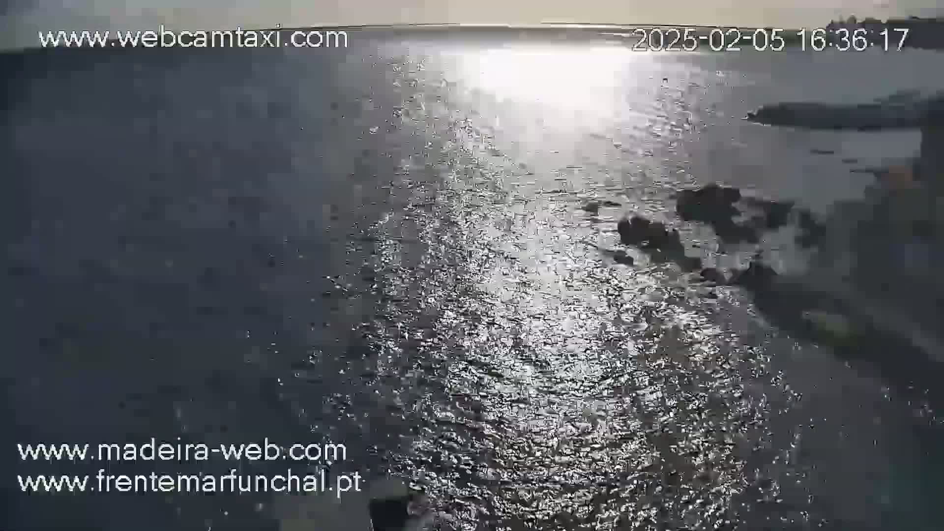 A high-angle view shows a body of dark water reflecting sunlight, with dark rocks visible near the shore under a mostly clear sky.