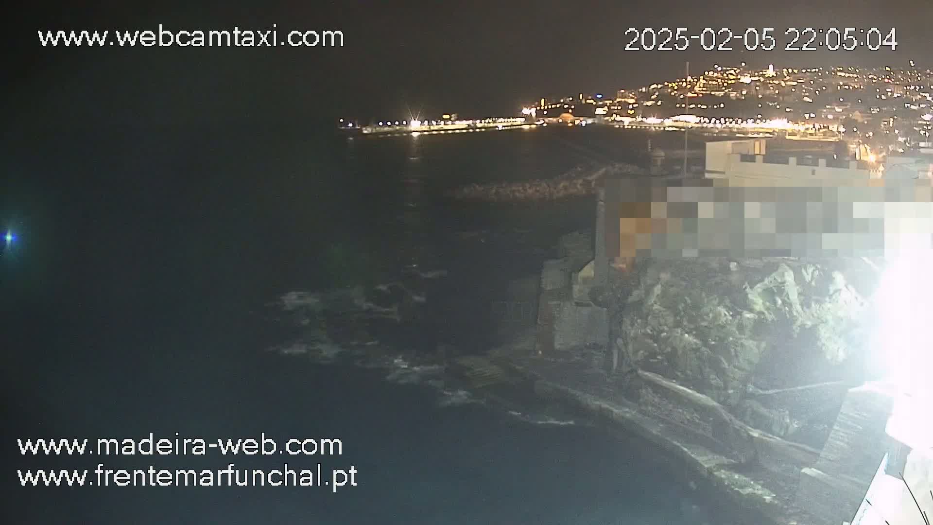 A nighttime coastal view shows a dark, choppy ocean with a city's lights in the distance and a rocky shoreline with a building partially obscured.