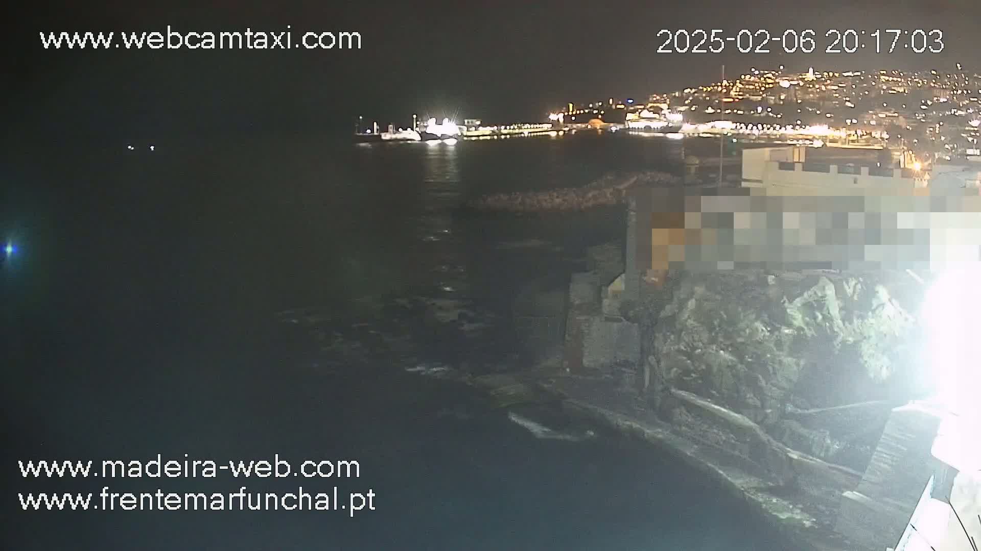 A nighttime view of a dark body of water with a city brightly lit on the opposite shore, a rocky structure in the foreground, and a ship visible in the distance.