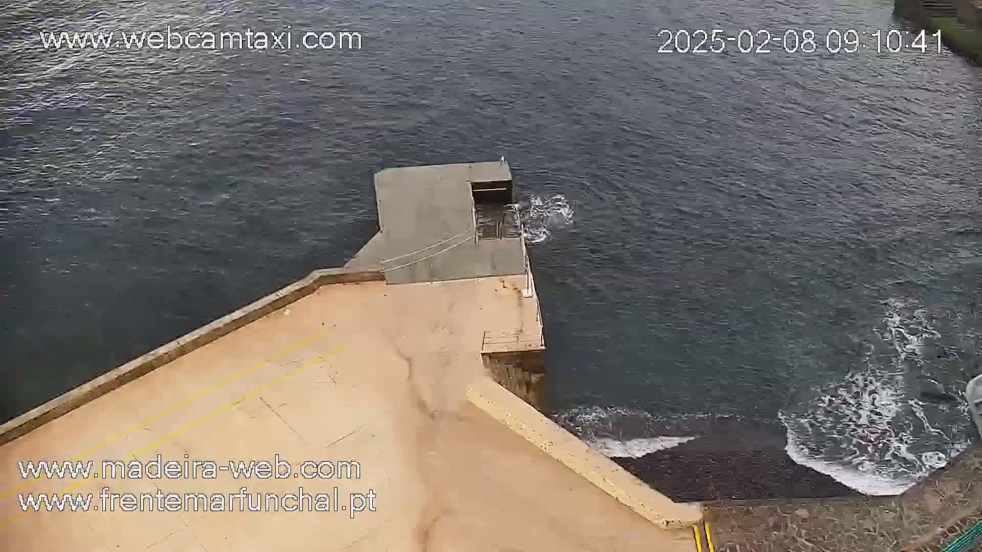An aerial view shows a concrete platform extending from a stone shoreline into dark, slightly choppy water.