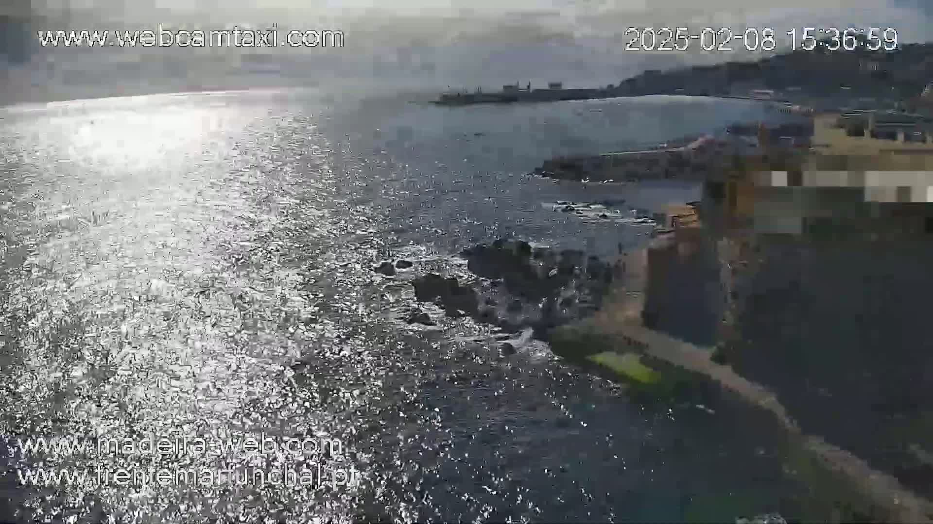A partly sunny day shows a rocky coastline with shimmering water, and a town visible in the distance.