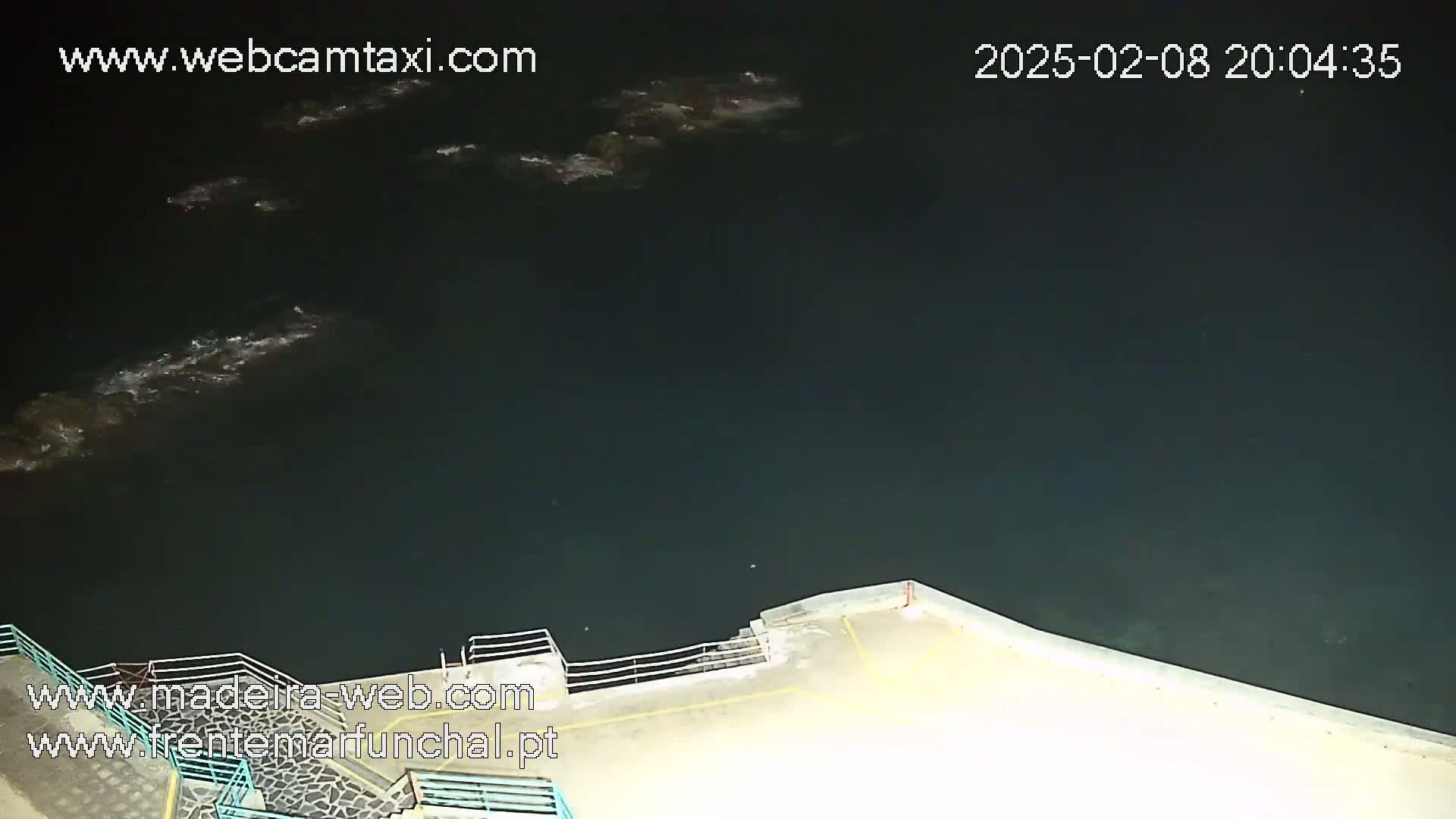 A nighttime, high-angle view shows dark water with small waves near a cement waterfront with railings.