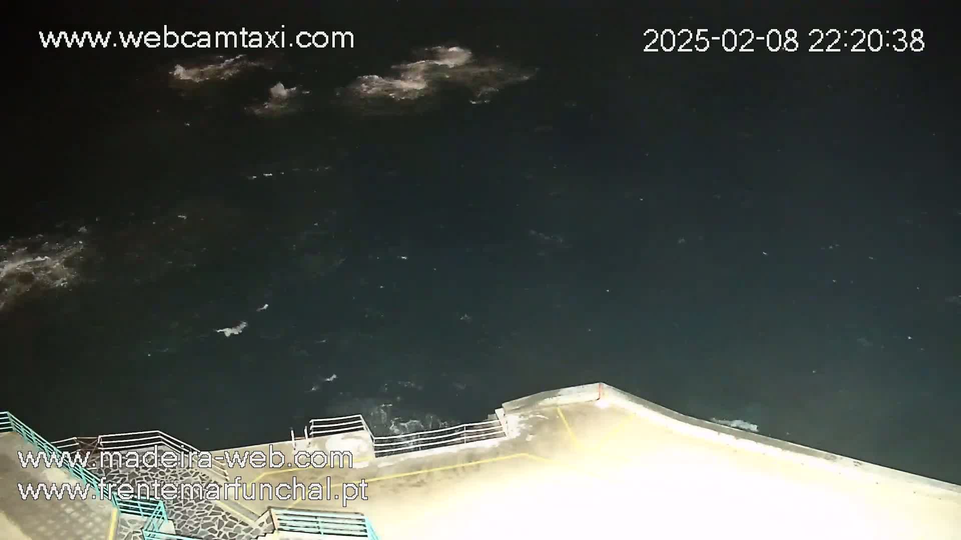 A nighttime, high-angle view shows dark water with some whitecaps near a low concrete wall with a railing.