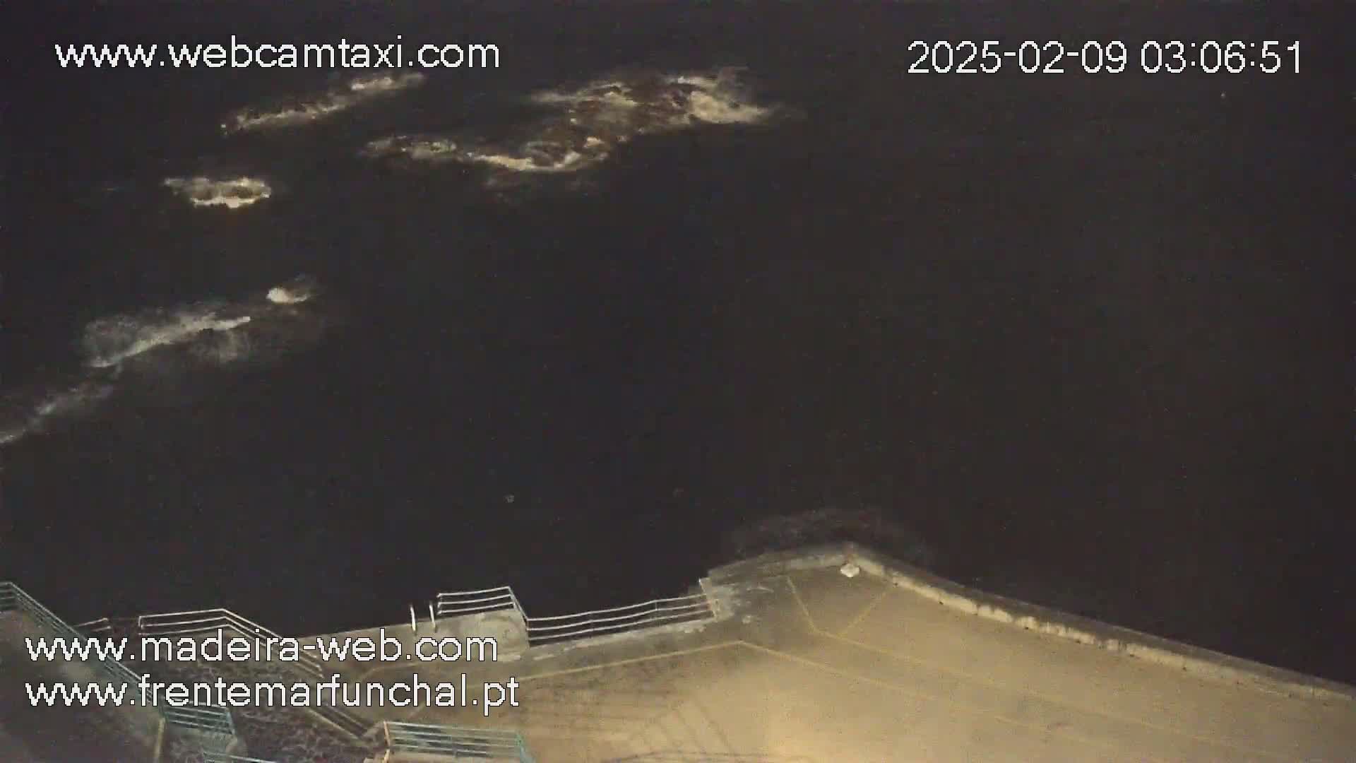 A nighttime, high-angle view shows dark water with small areas of white foam, a concrete embankment, and a railing.