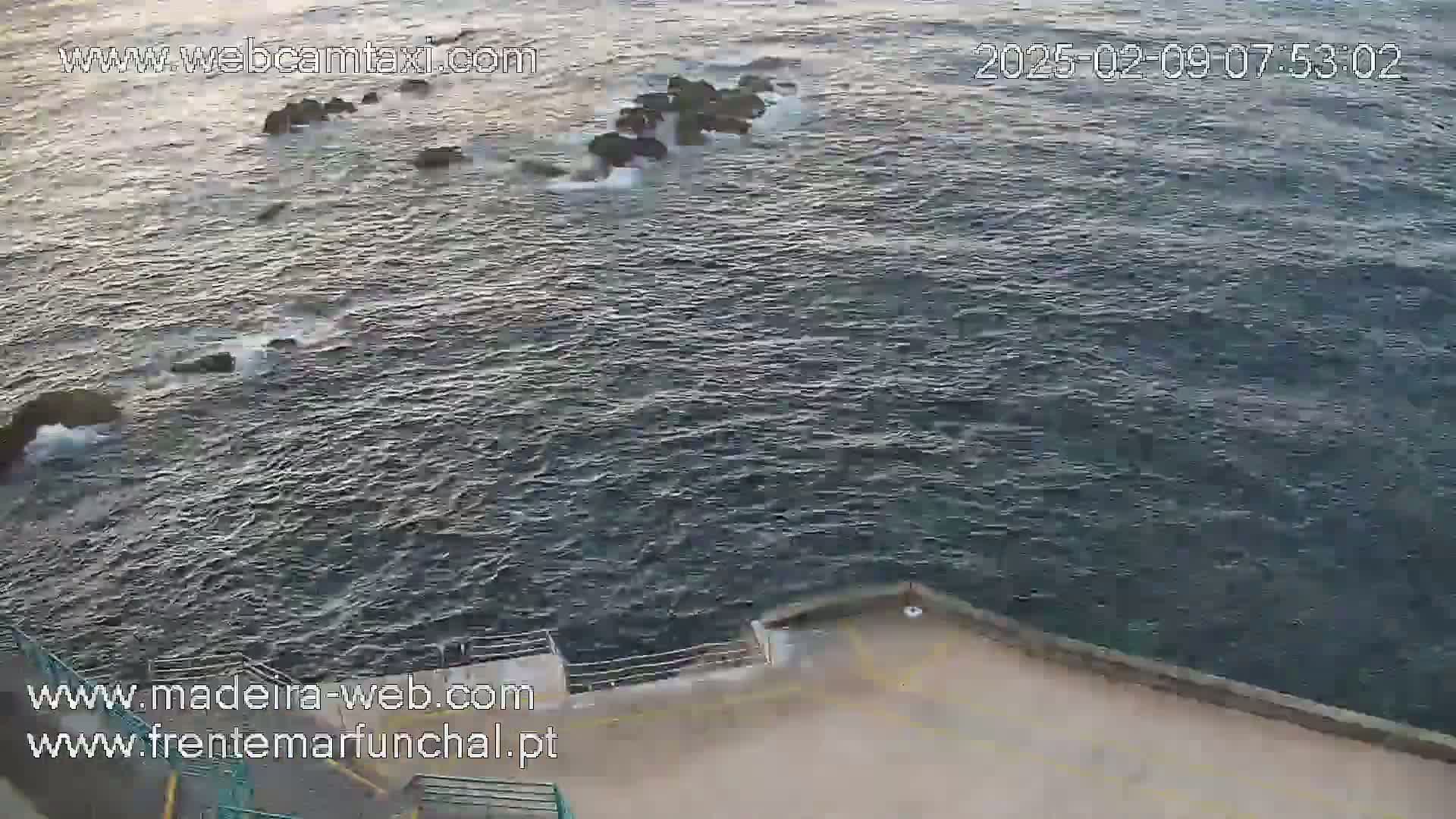 A high-angle view shows dark, choppy ocean water lapping against a concrete wall on a somewhat overcast day.