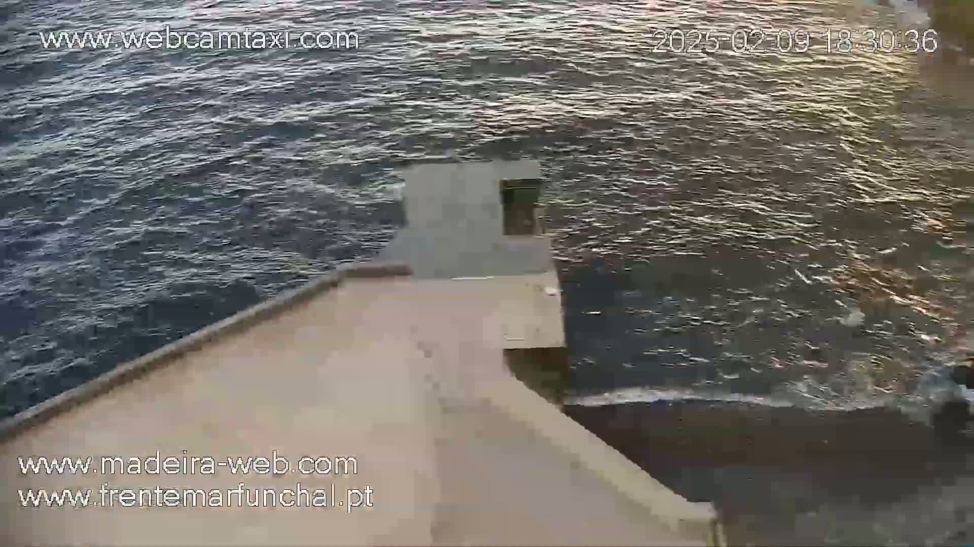 A high-angle view shows a dark-colored body of water with small waves lapping against a light-beige structure, under what appears to be somewhat overcast conditions.