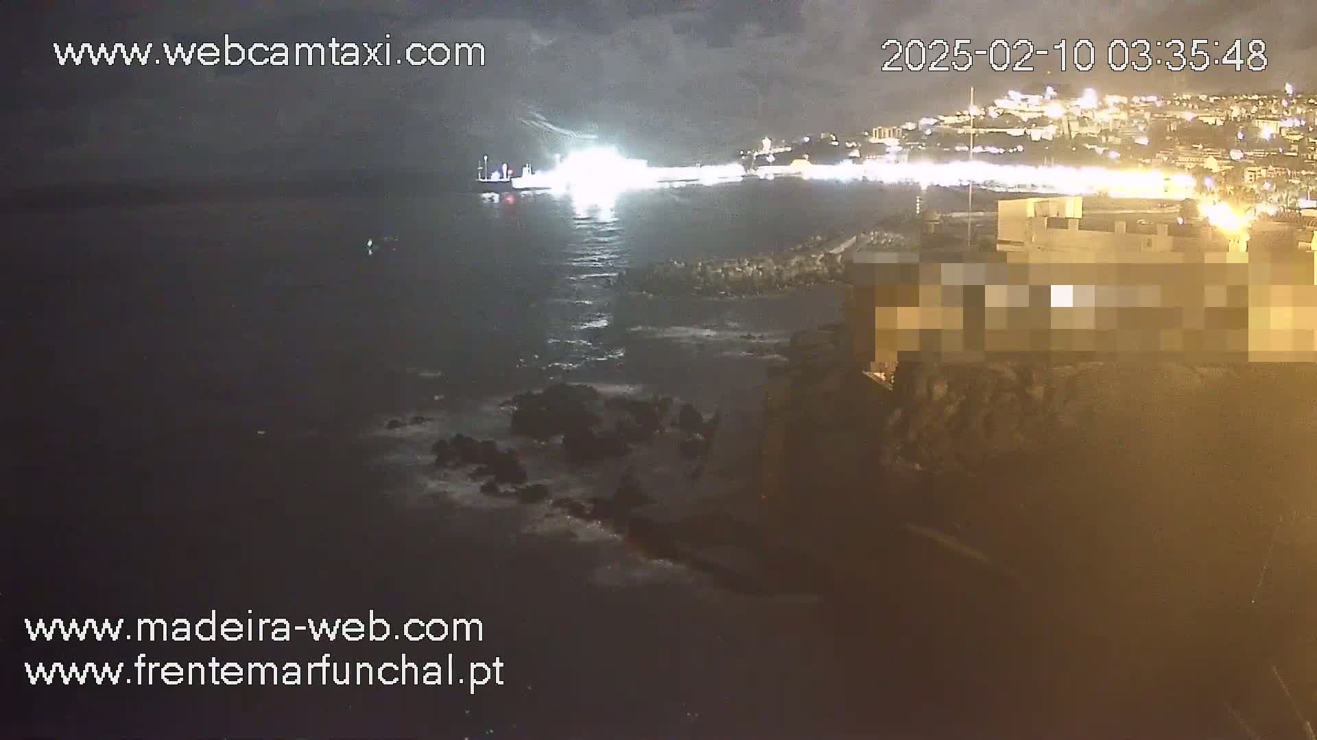 A nighttime, coastal scene shows a body of dark water with a ship and city lights in the distance, along with a rocky shoreline and a building.