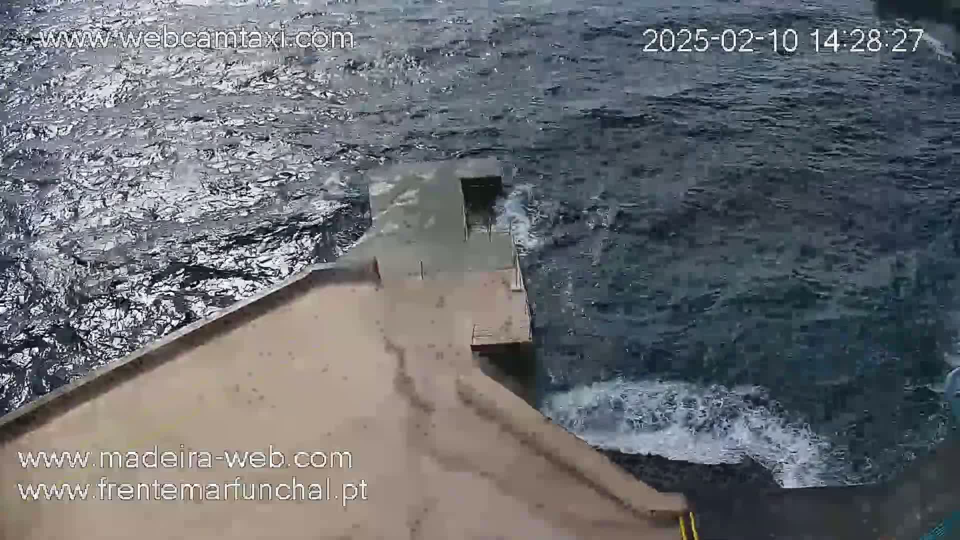 An aerial view shows a concrete platform extending into dark, choppy ocean water with small waves breaking against it.