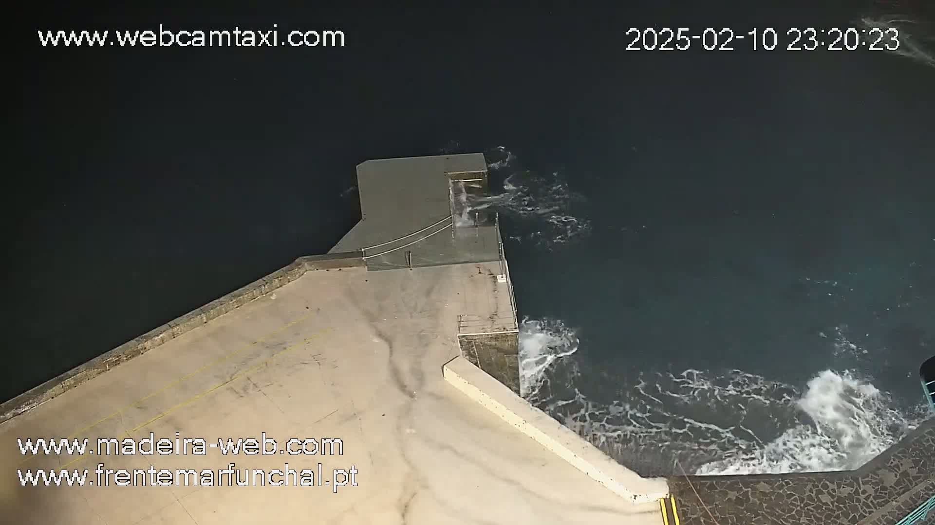 A high-angle nighttime view shows waves crashing against a concrete structure extending into dark water.