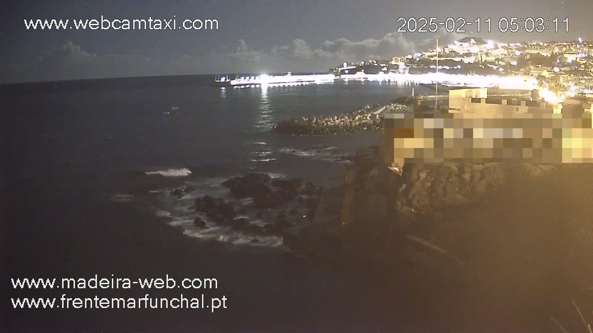 A nighttime coastal scene shows dark water lapping against rocky shores, with a brightly lit city in the background under a partly cloudy sky.