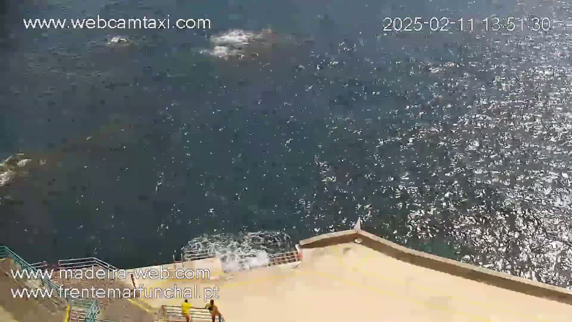 A high-angle view shows a choppy ocean meeting a tan wall with two people near the bottom of some stairs.