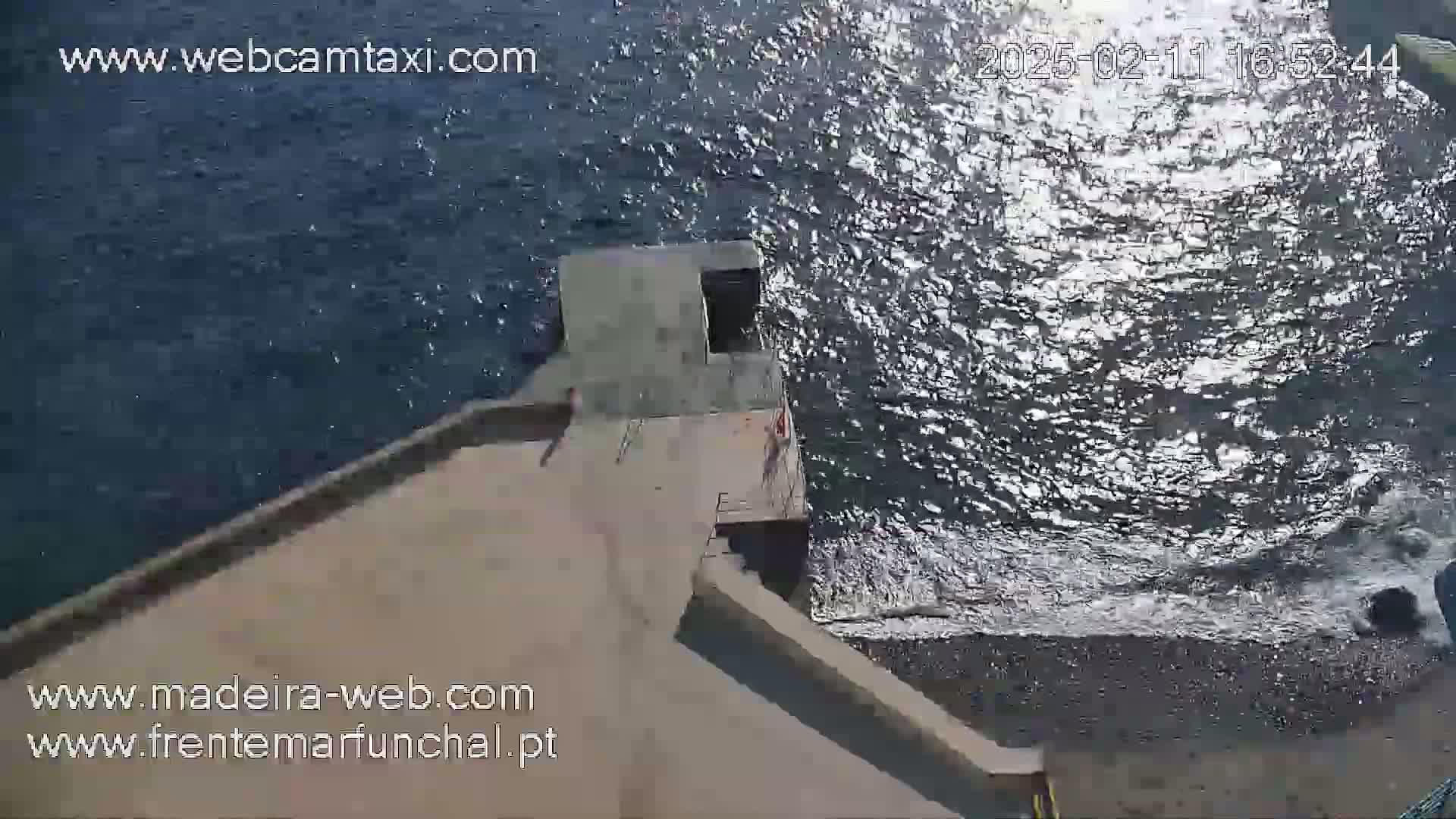 An aerial view shows a cement platform extending from a shoreline into a body of dark, glistening water on a sunny day.