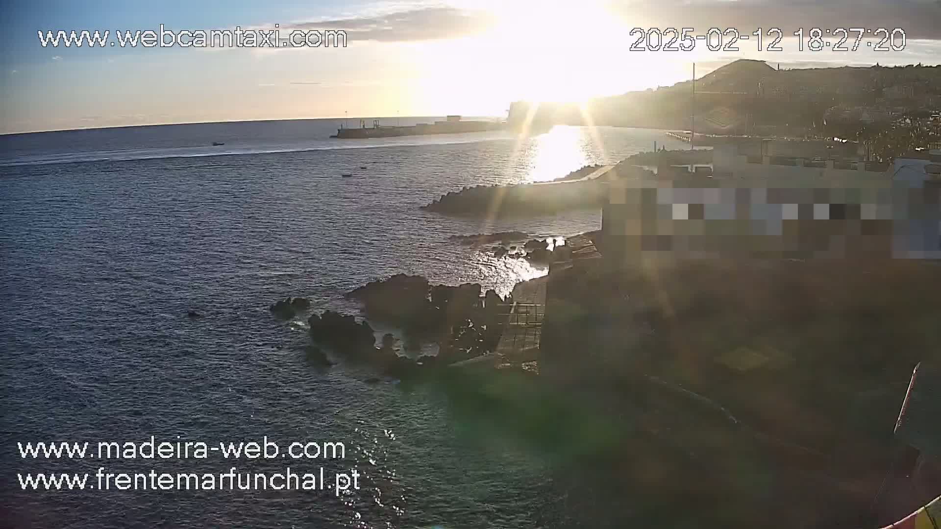 A sunlit ocean scene shows a rocky shoreline with buildings and a distant town under a mostly clear sky.