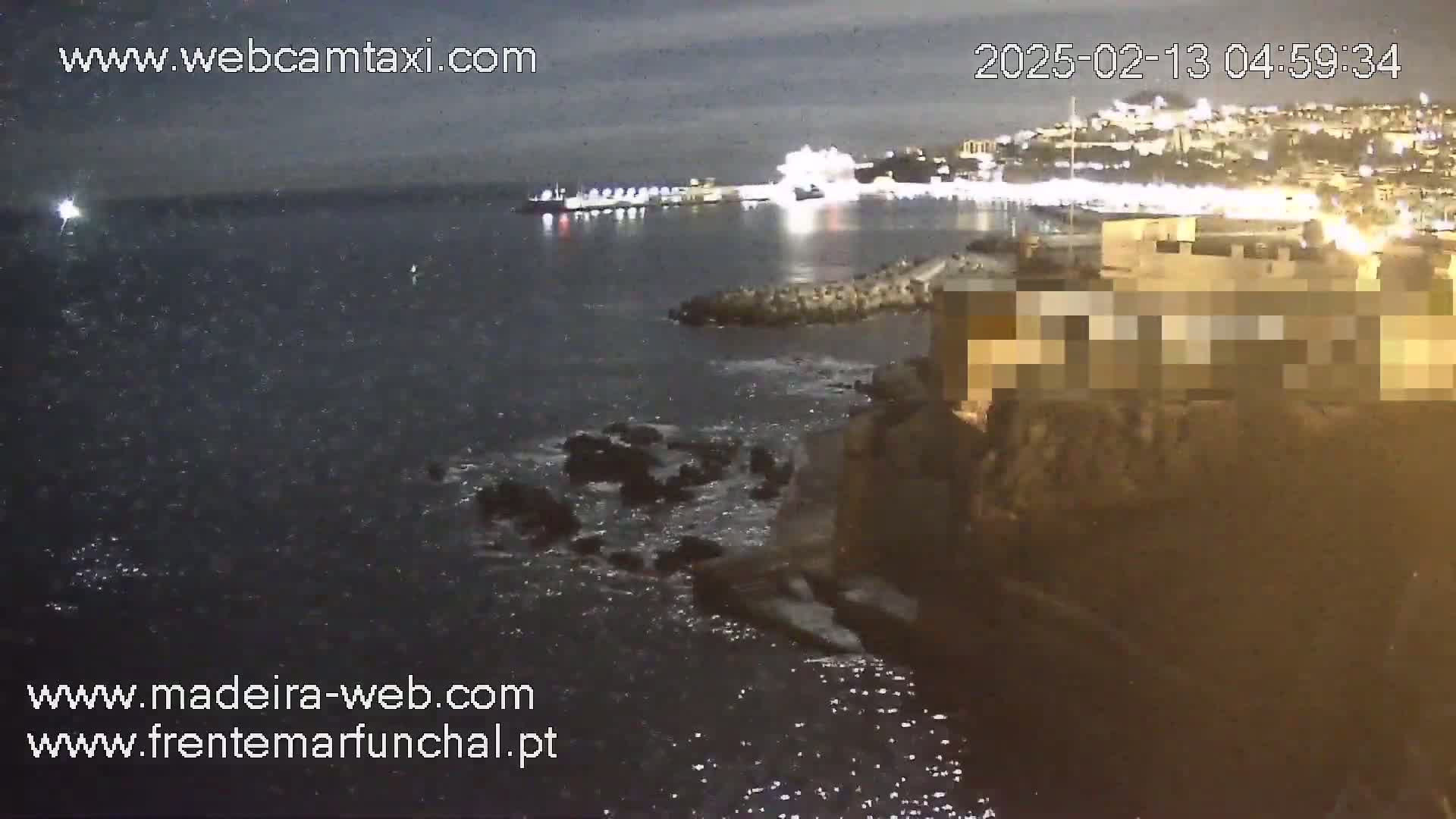A nighttime view of a dark, wavy ocean with rocky shoreline and a brightly lit city in the distance under a cloudy sky.