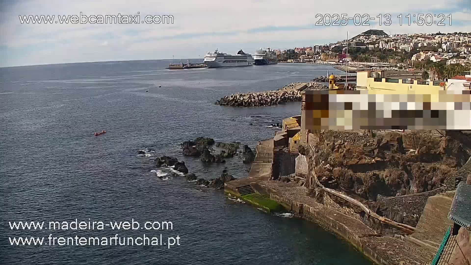 A partly sunny day shows two cruise ships in a harbor next to a rocky coastline with a small building and a few people kayaking in the ocean.
