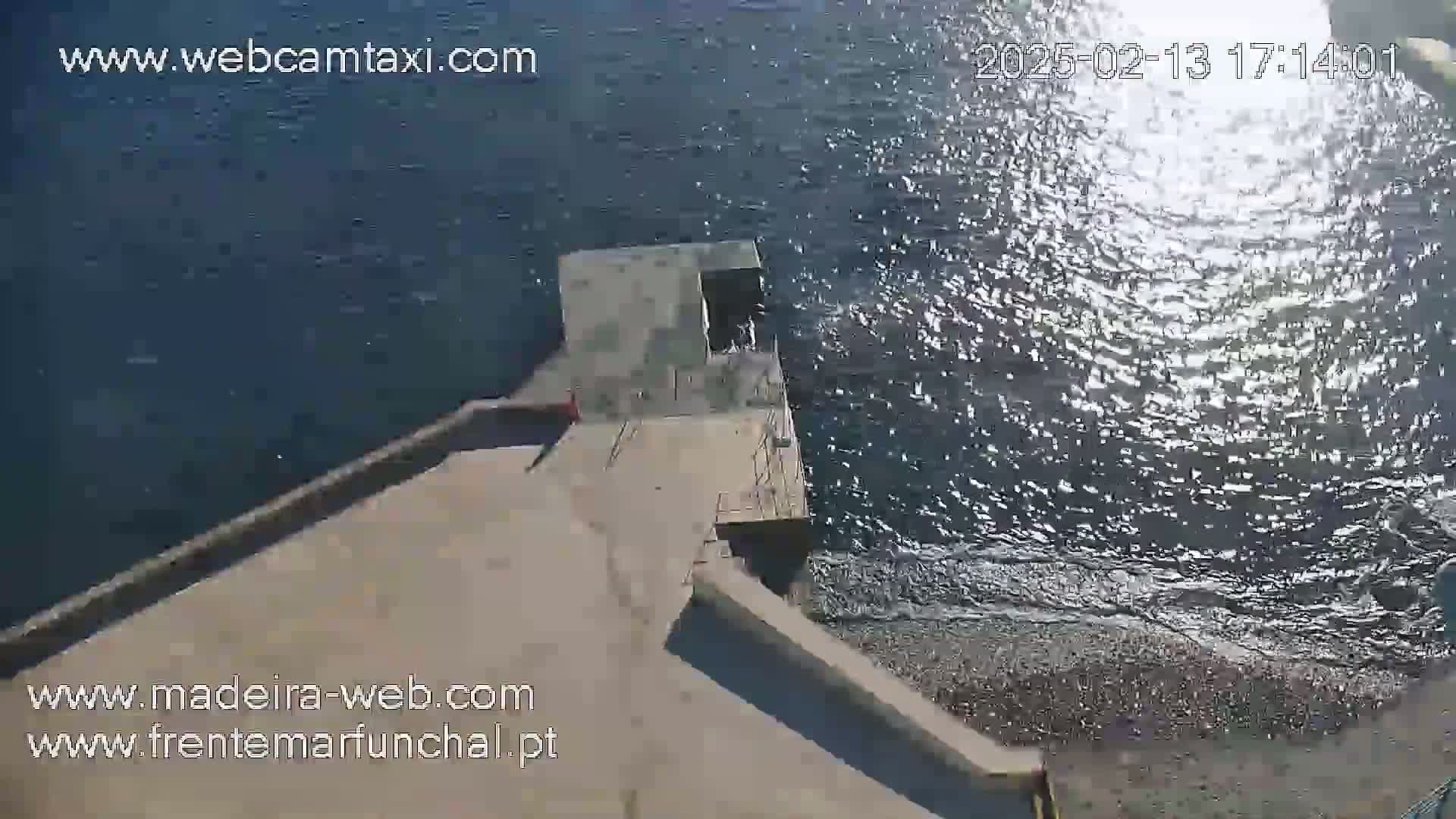 An aerial view shows a concrete platform extending from a shoreline into a calm, sunlit body of water.