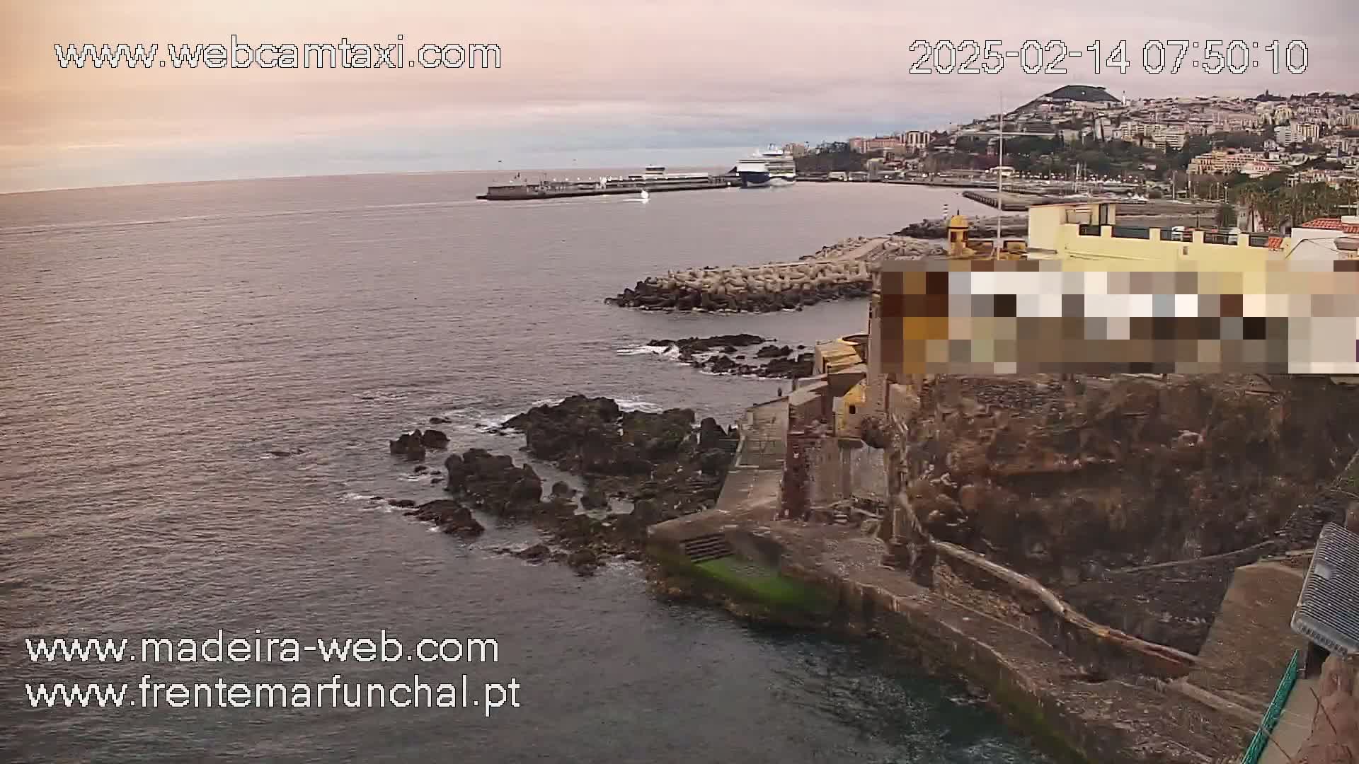 A large body of calm water laps against rocky shores near a city under an overcast sky.