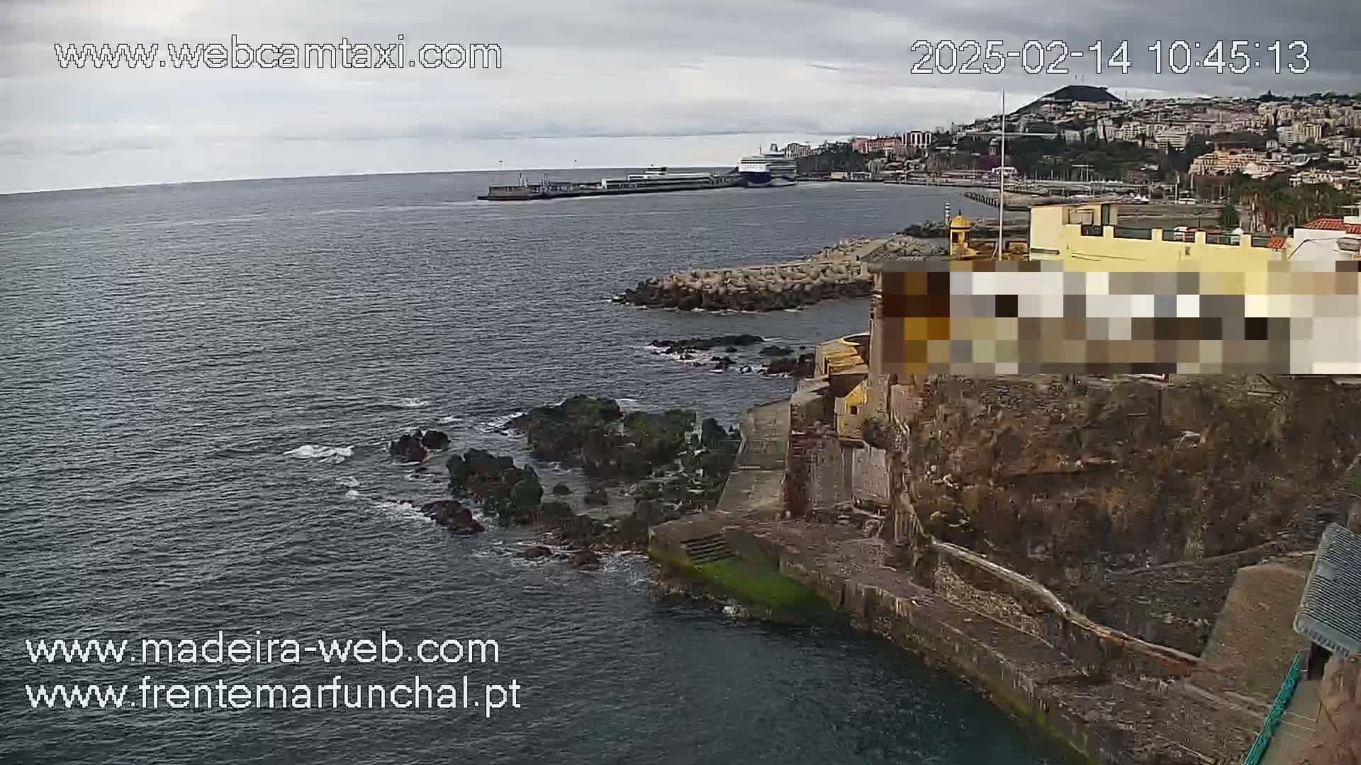 A large ferry is docked in a harbor, with a rocky coast and a partially obscured building on the right, under an overcast sky.