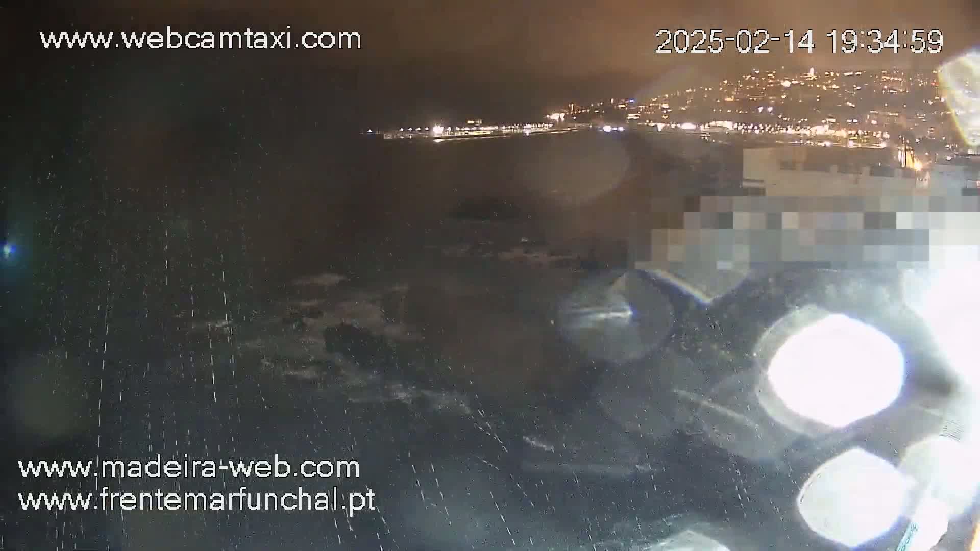 A nighttime view shows rain falling over dark ocean waves crashing against a shoreline with a brightly lit city in the background.