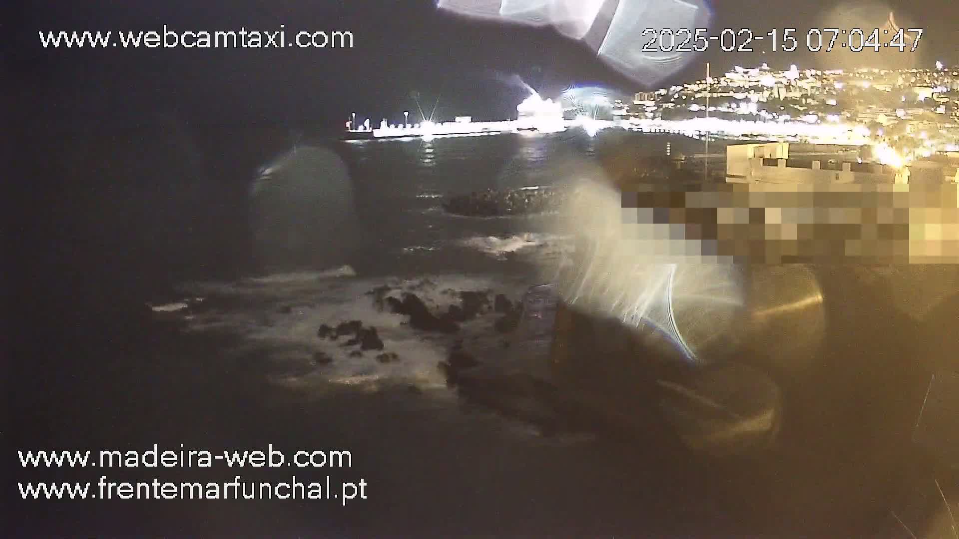 A nighttime coastal scene shows waves crashing against rocks near a brightly lit city under a dark sky.