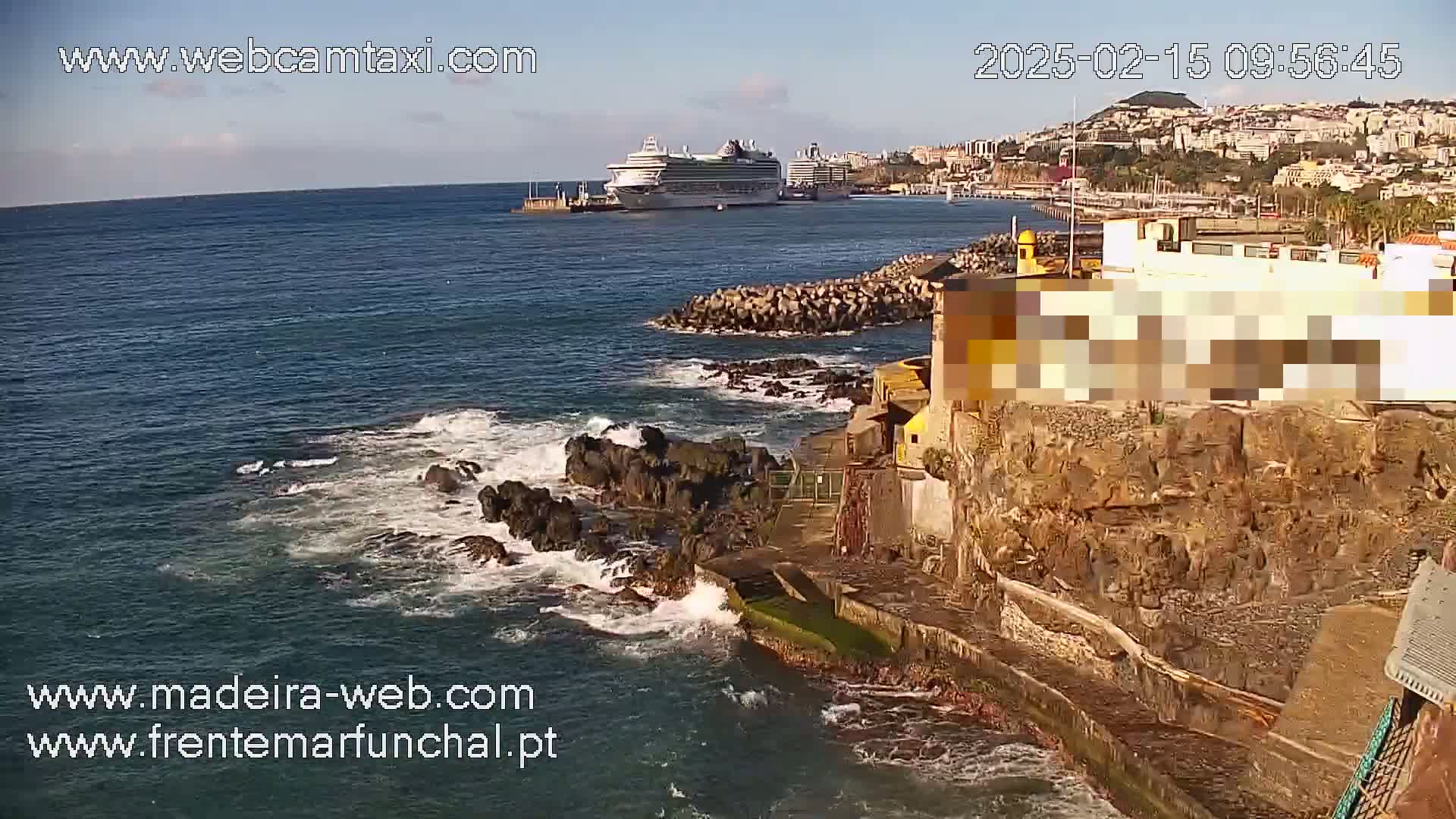 A cruise ship is visible in the distance of a clear, sunny day, as waves crash against dark rocky shores near a building.
