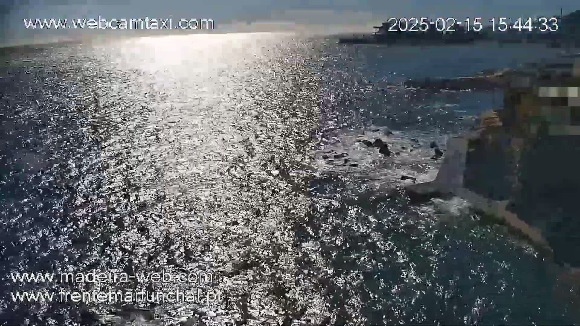 A body of water with waves and sunlight reflecting on its surface, near a rocky shoreline on a partly sunny day.