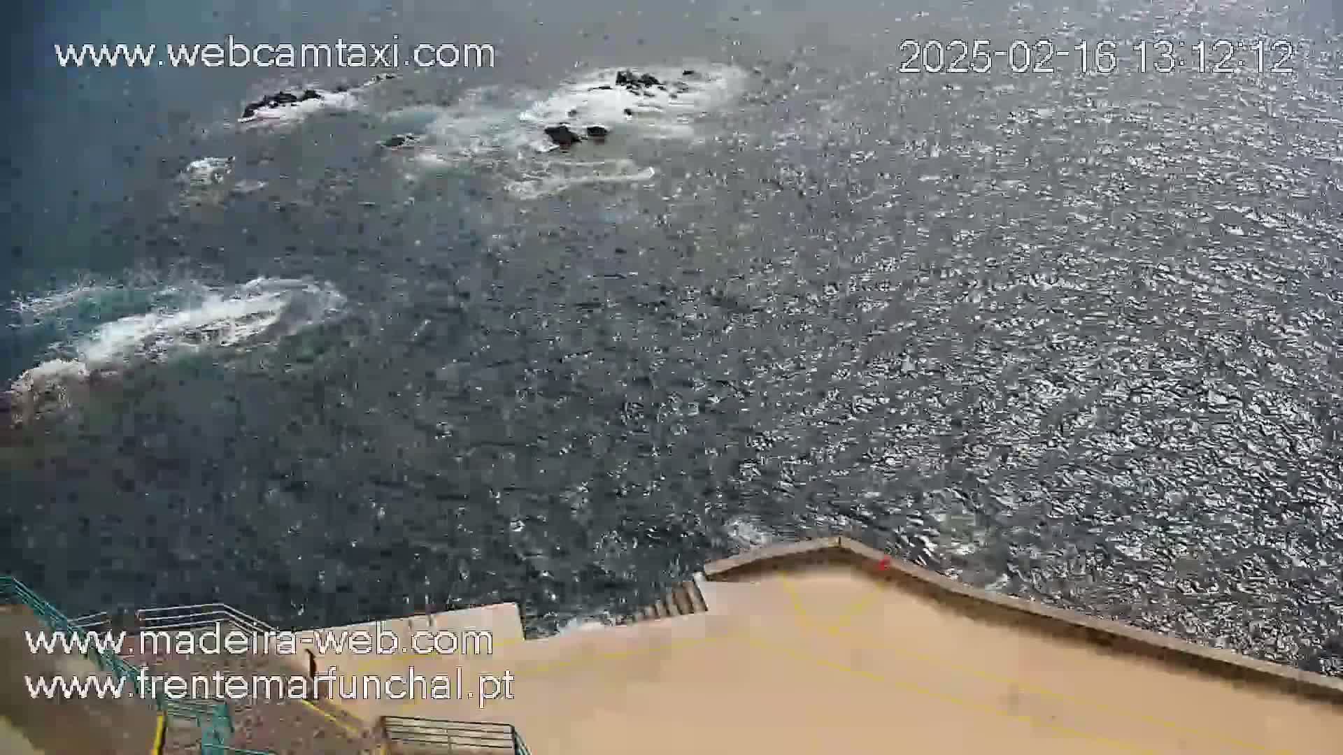 A high-angle view shows dark, choppy ocean water lapping against a tan, paved shoreline with steps leading down to the water, and a few dark rocks visible in the water.