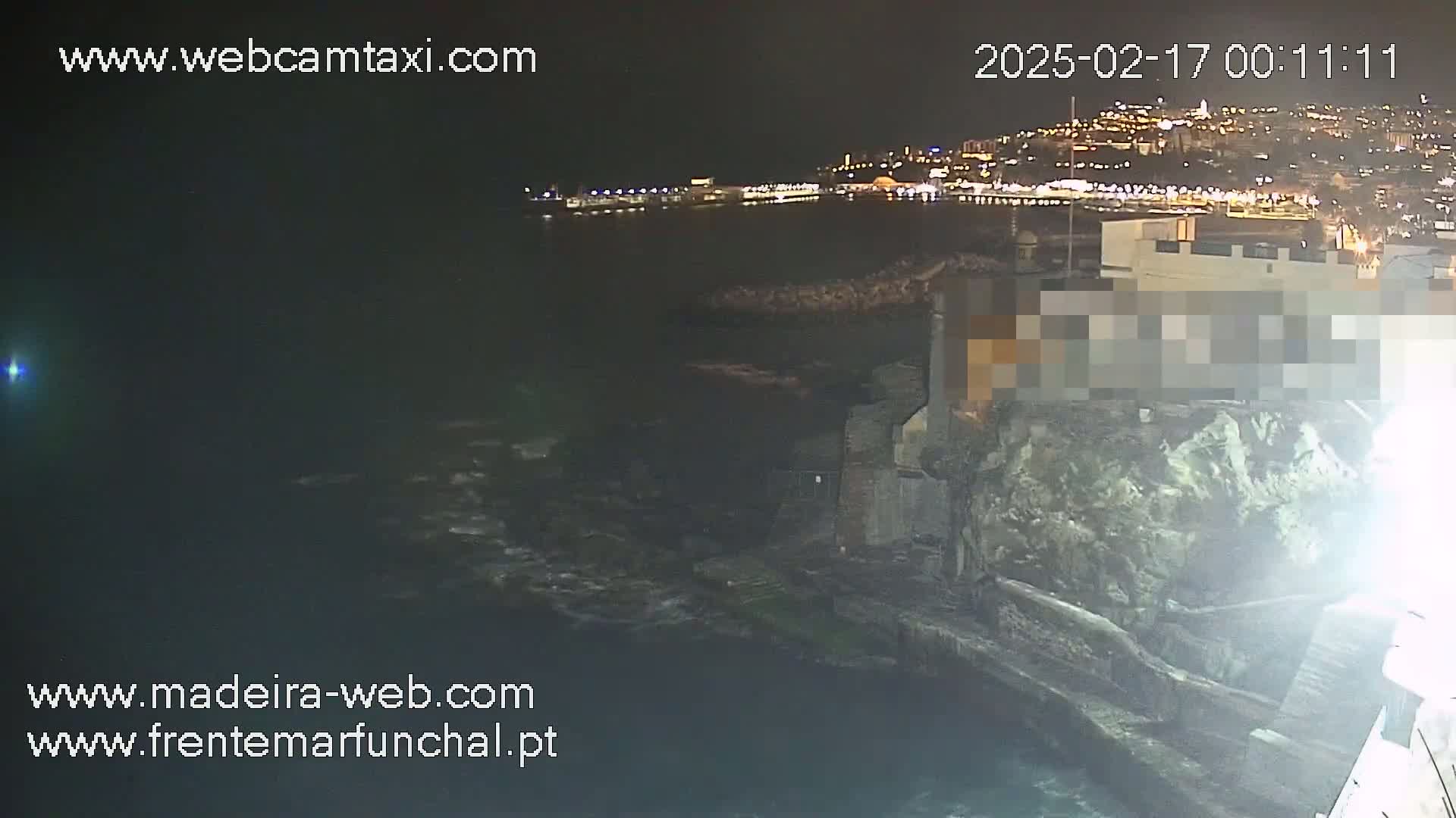 A nighttime coastal scene shows a dark body of water with some wave action, a rocky shoreline with buildings, and a brightly lit city in the distance.