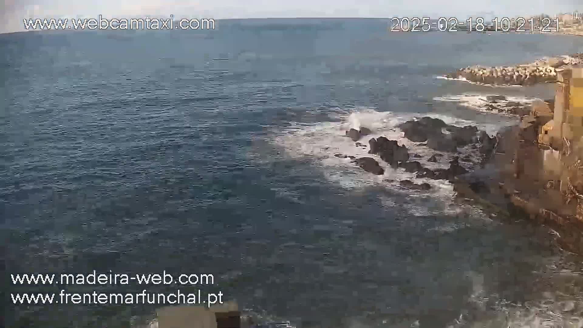 Ocean waves crash against dark rocks near a stone building on a partly cloudy day.