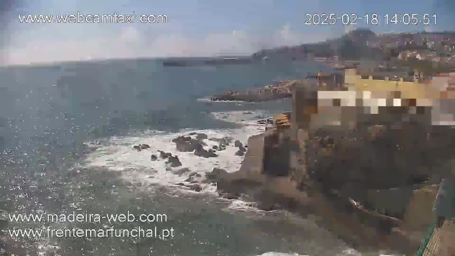 Ocean waves crash against a rocky shoreline near a coastal town on a partly cloudy day.
