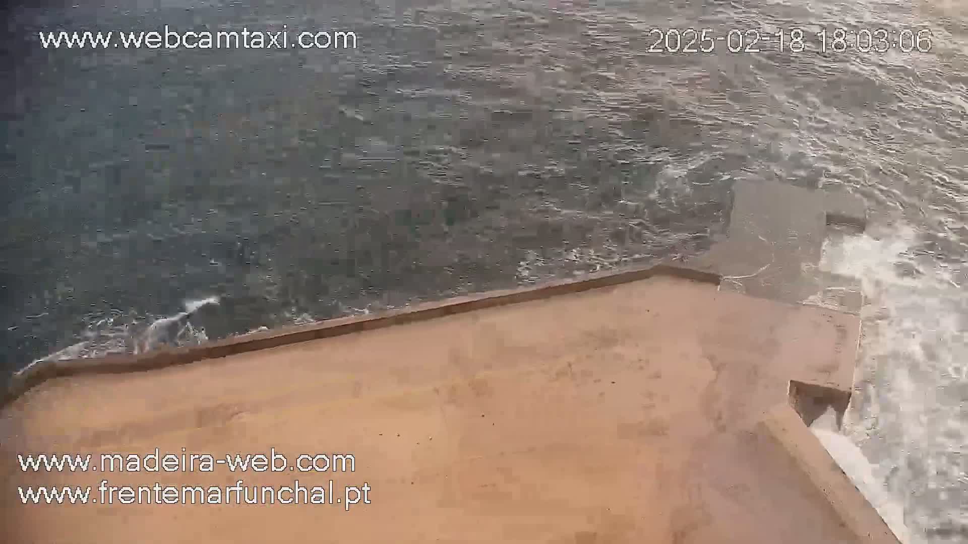 A high-angle view shows choppy waves crashing against a tan seawall.