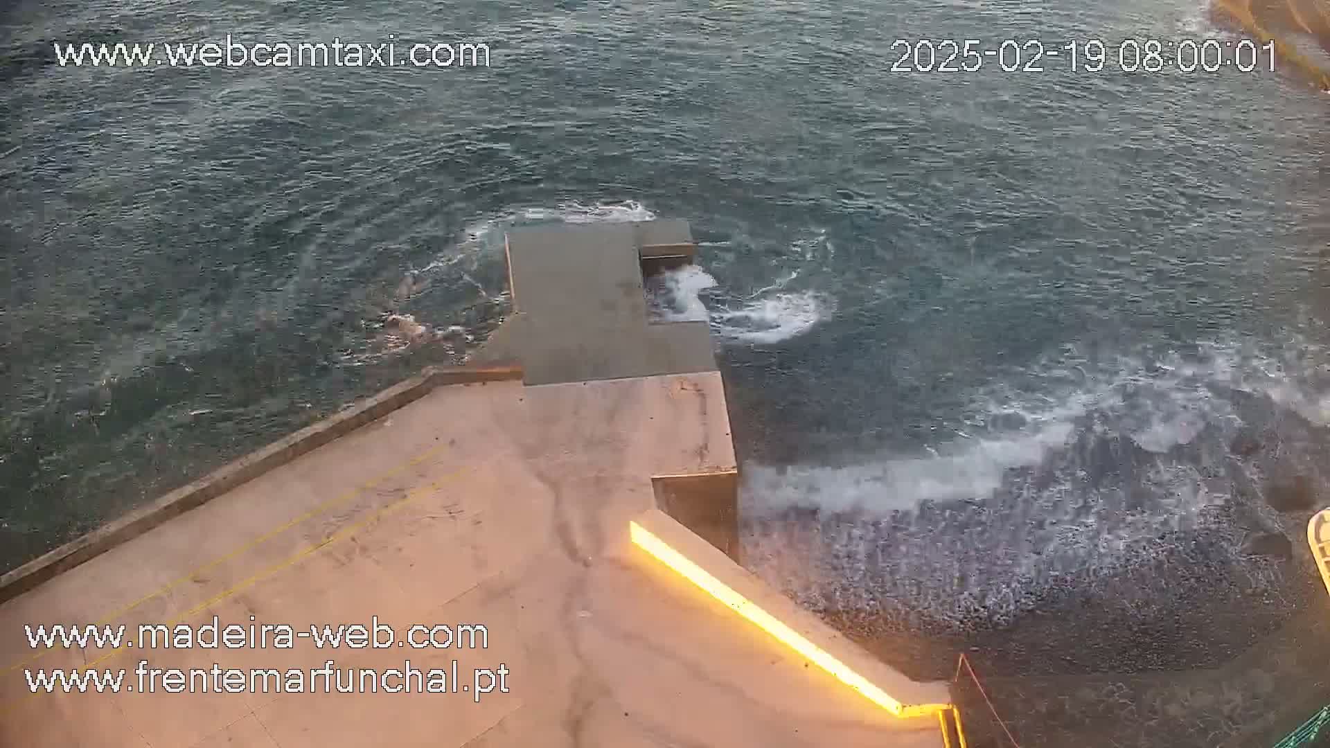 A high-angle view shows a concrete structure near a choppy sea with waves crashing against the rocks.