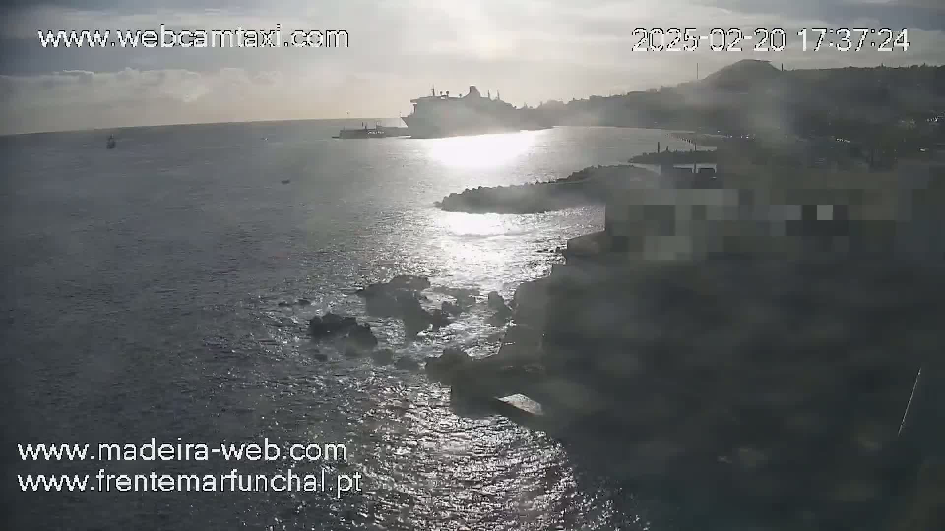 A large cruise ship is docked near a rocky coastline under a partly cloudy sky, with sunlight reflecting on the water.